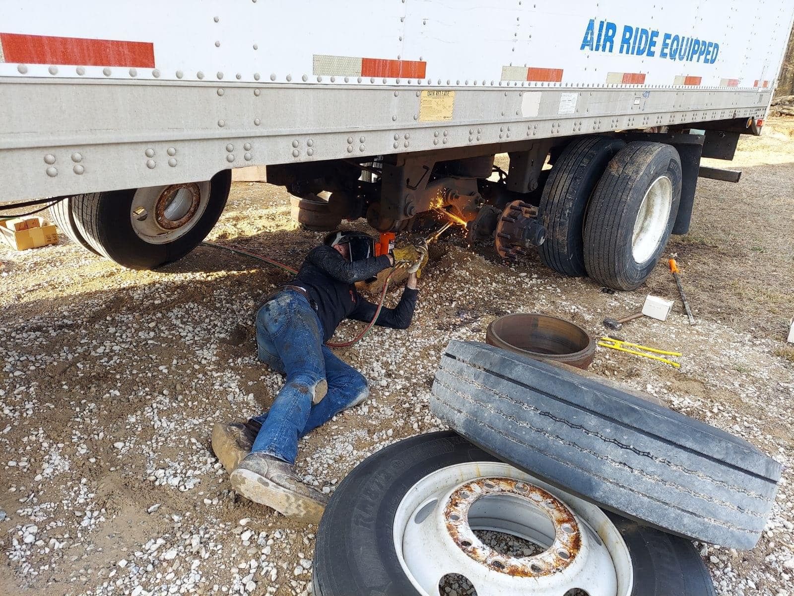 A person welding under a semi-truck trailer, using a torch. They are wearing safety gear.