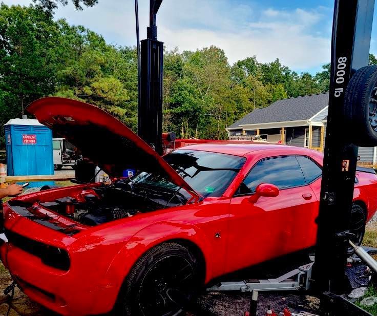 Red Dodge Challenger on a car lift with the hood open, outdoors, next to a portable toilet and shed.