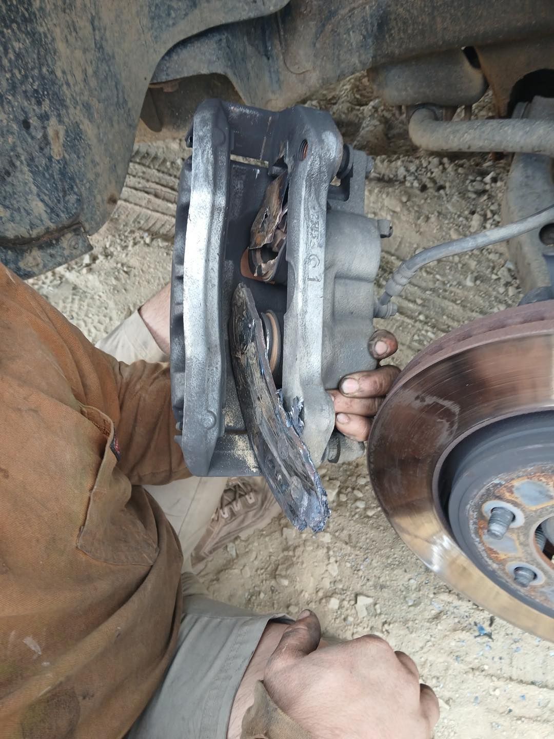Brake caliper detached from rotor held by a person. Outdoors, rusted components, worn brown shirt.