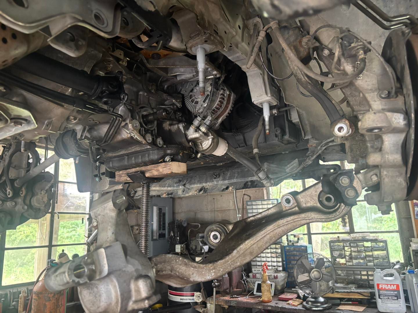 Undercarriage of a vehicle undergoing repair, showing suspension components, exposed engine, and tools in a garage setting.