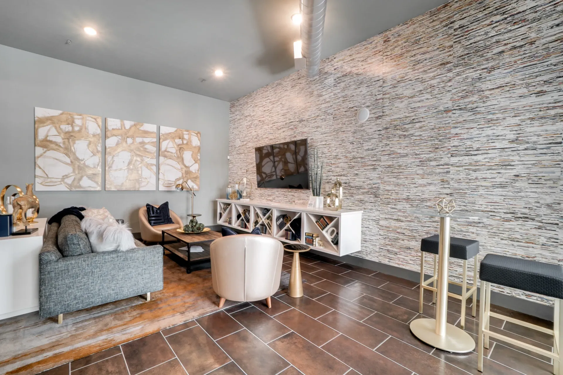 Modern resident lounge with grey sofa, beige chairs, and a TV on a textured wall.