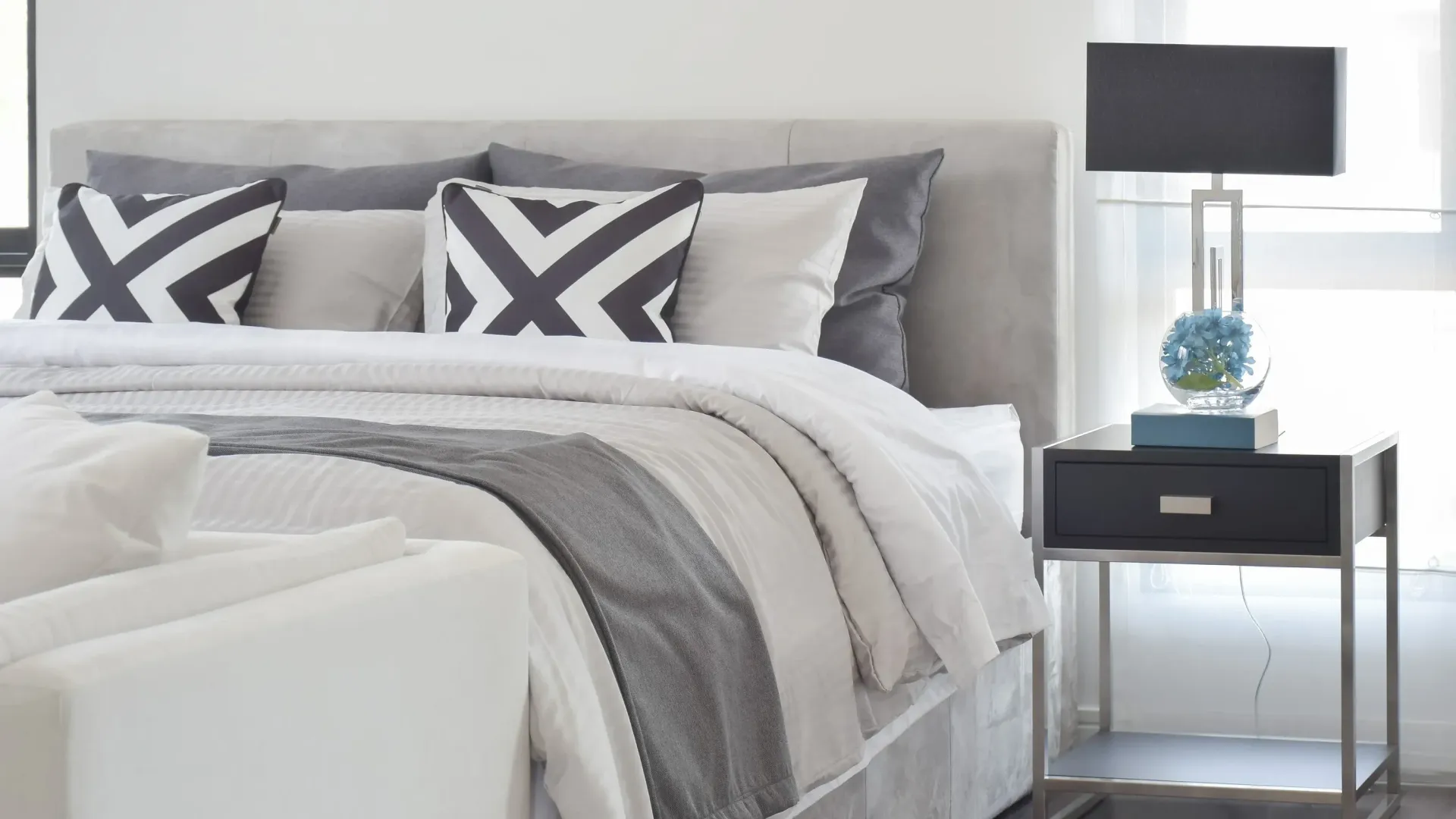 Bedroom with gray and white bedding, decorative pillows, and a modern nightstand with a lamp.