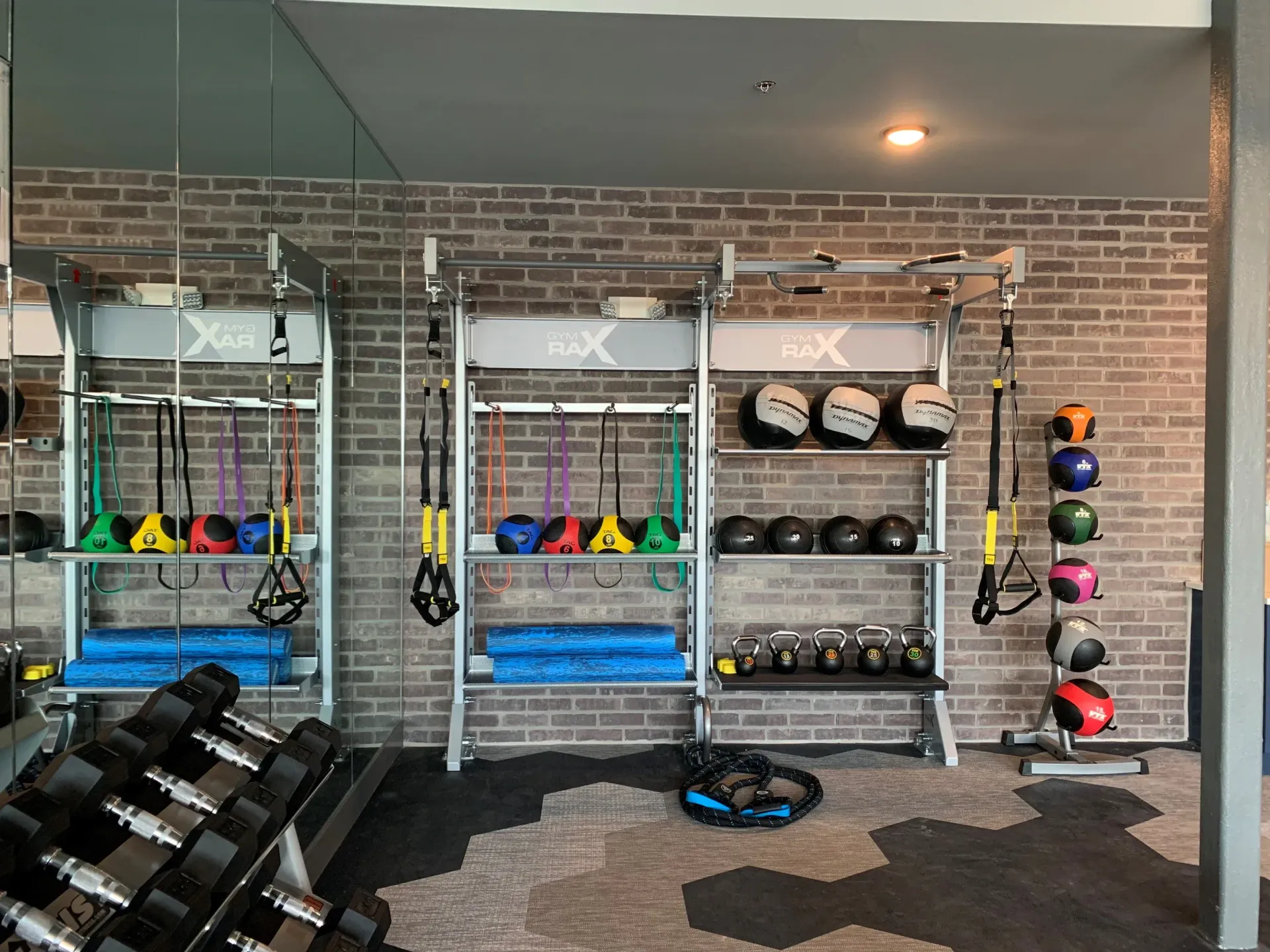 Gym area with weight racks, medicine balls, kettlebells, mats, and resistance bands against a brick wall.