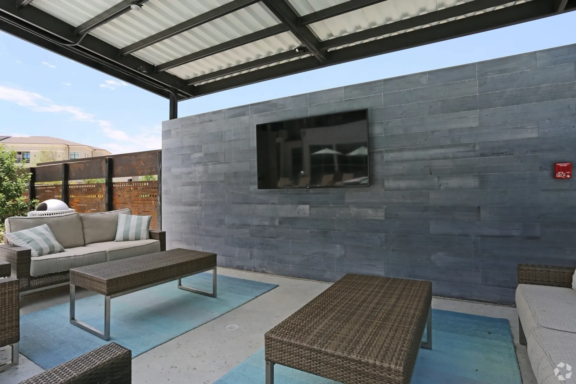 Outdoor community lounge with wicker seating, a coffee table, and a mounted TV on a gray wall.