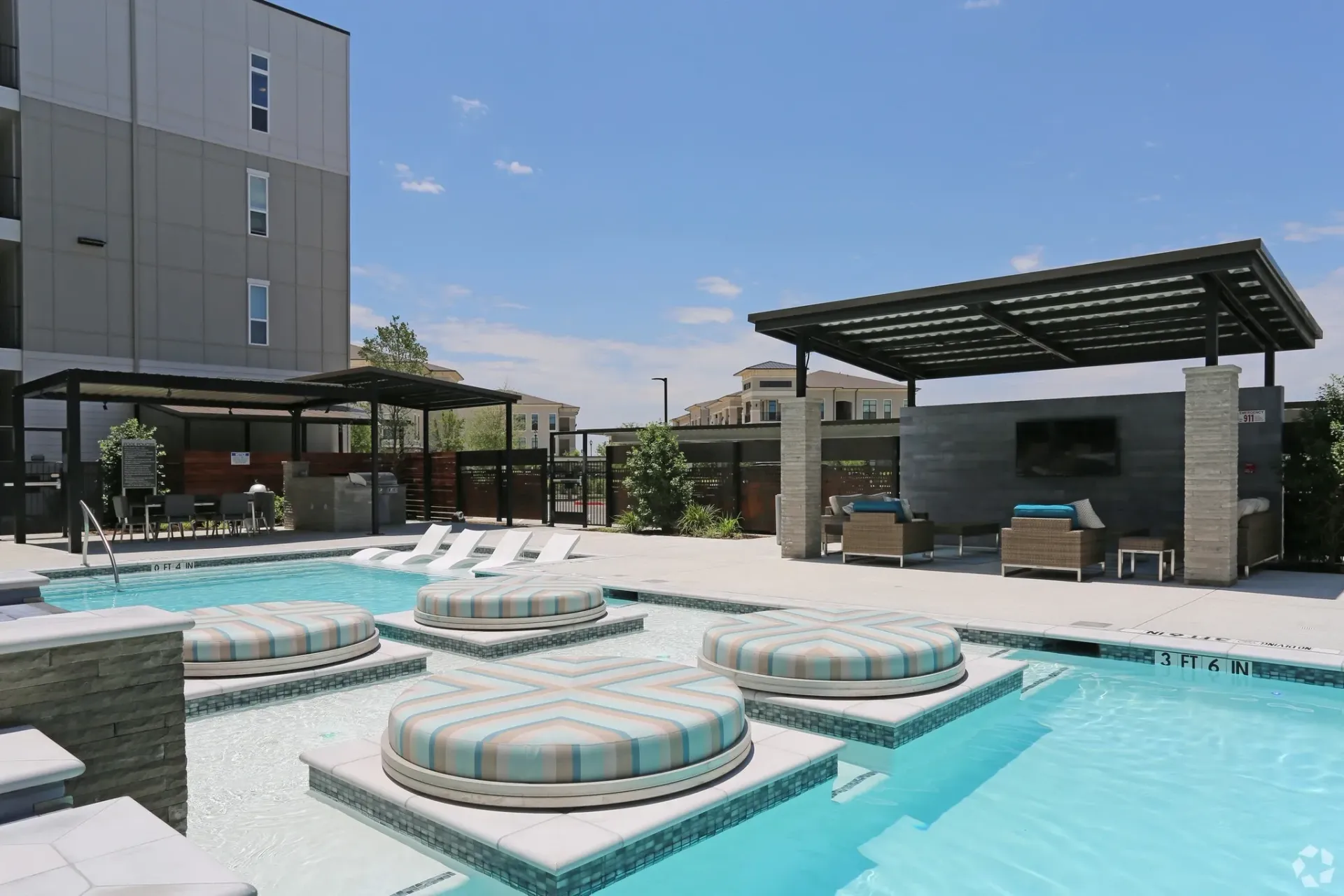 Outdoor community pool area with circular cushioned platforms, lounge chairs, and shaded seating.