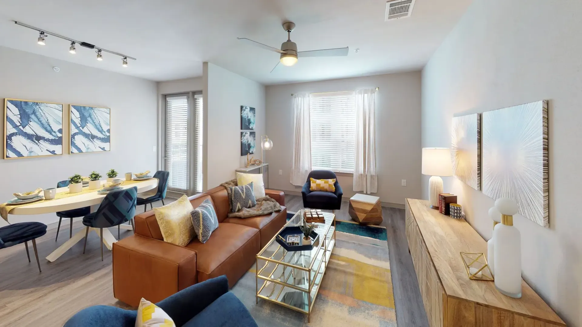 Open-concept living room in a contemporary apartment with a brown leather sofa, blue chairs, and a glass coffee table.