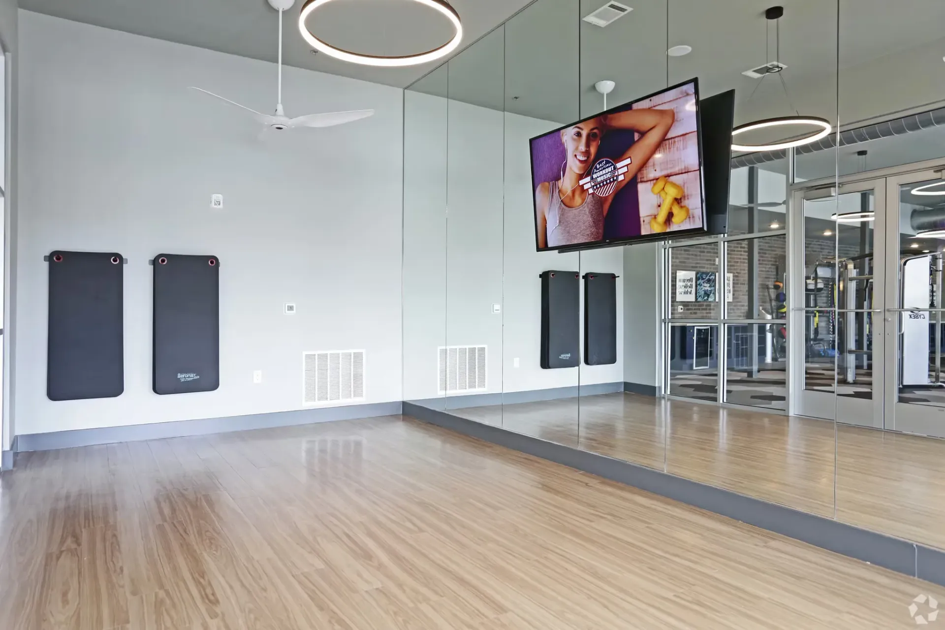 Fitness studio with mirrors, wood flooring, wall-mounted mats, and a TV.