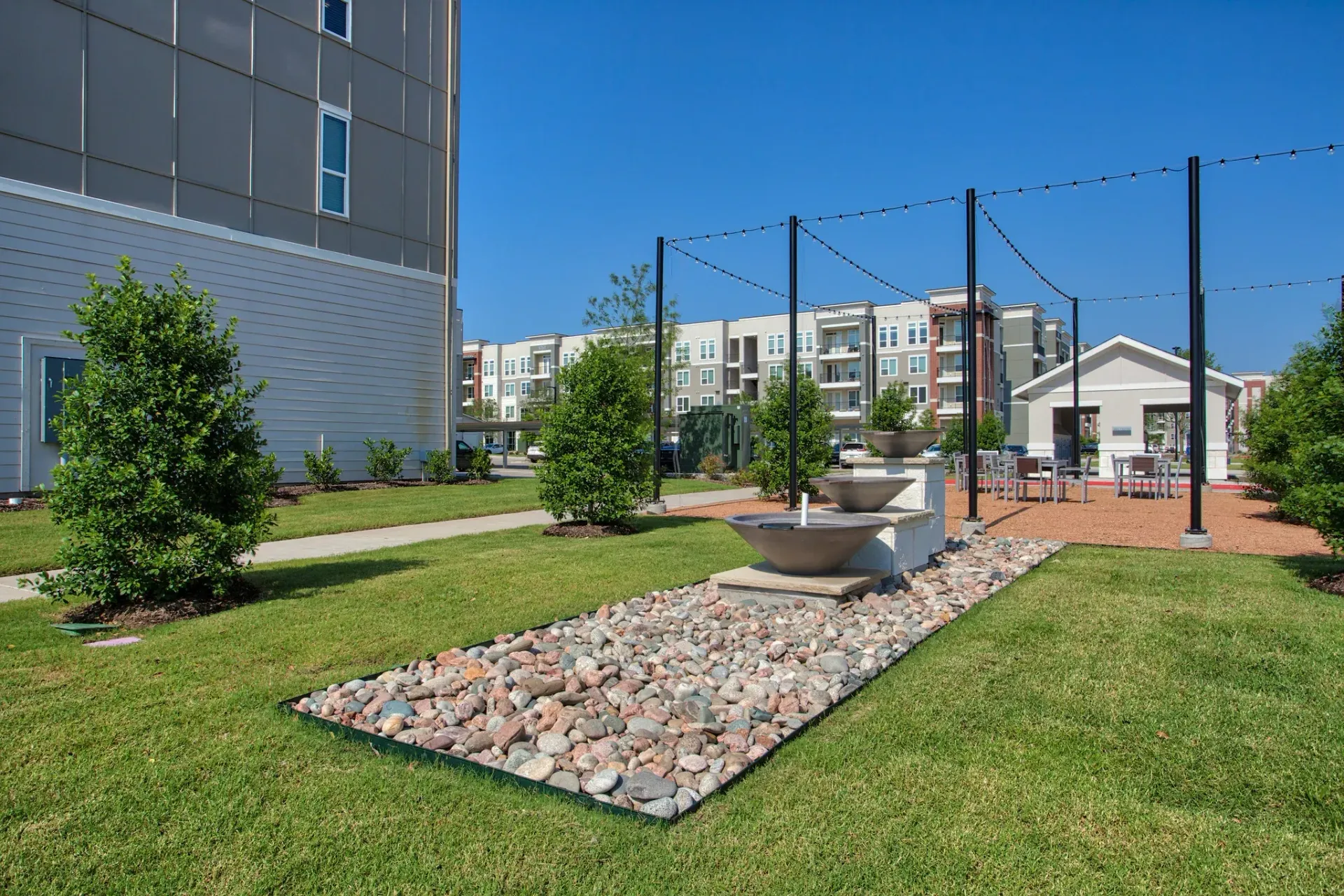 Outdoor community courtyard with green lawn, rock feature, string lights, and seating near apartment buildings.