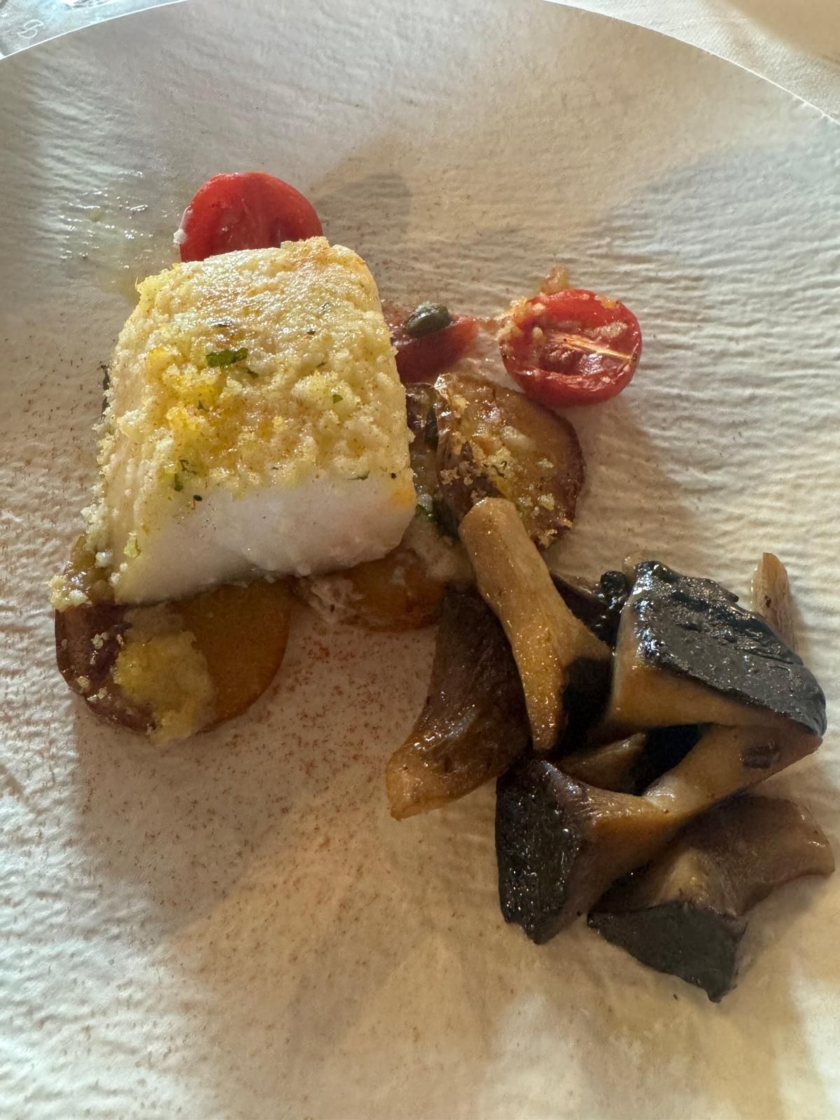 Piatto gourmet impiattato con filetto impanato, verdure arrostite, pomodorini e pezzi di funghi scuri su un piatto bianco.