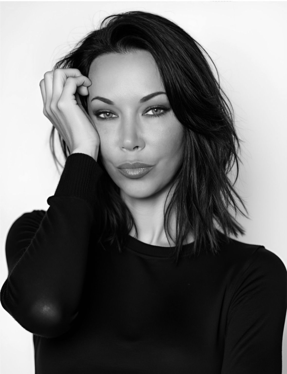 Woman with dark, wavy hair, hand on head, pouting lips, wearing a black long-sleeved shirt.