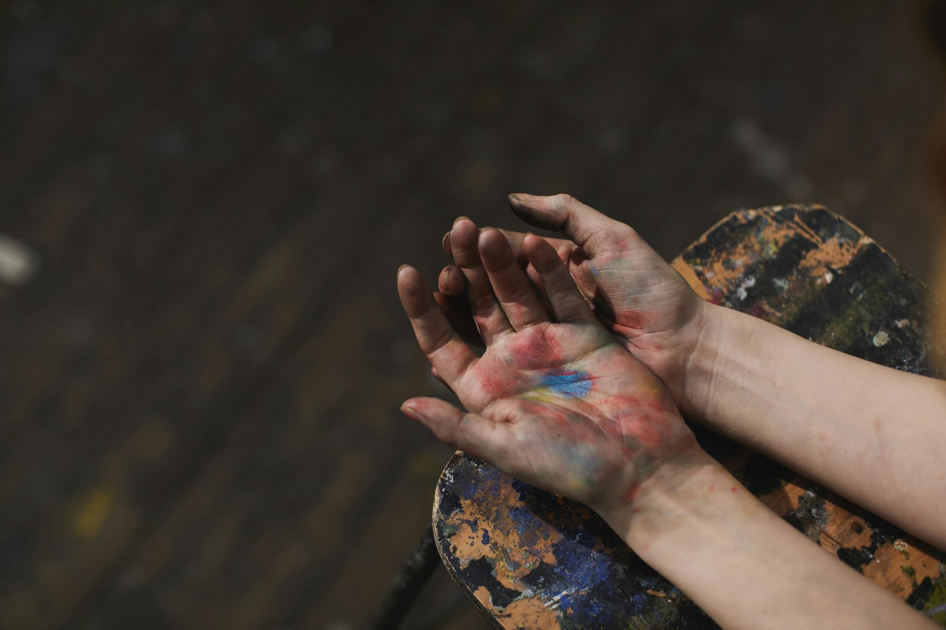 Hands covered in colorful paint, resting on a paint-splattered surface.