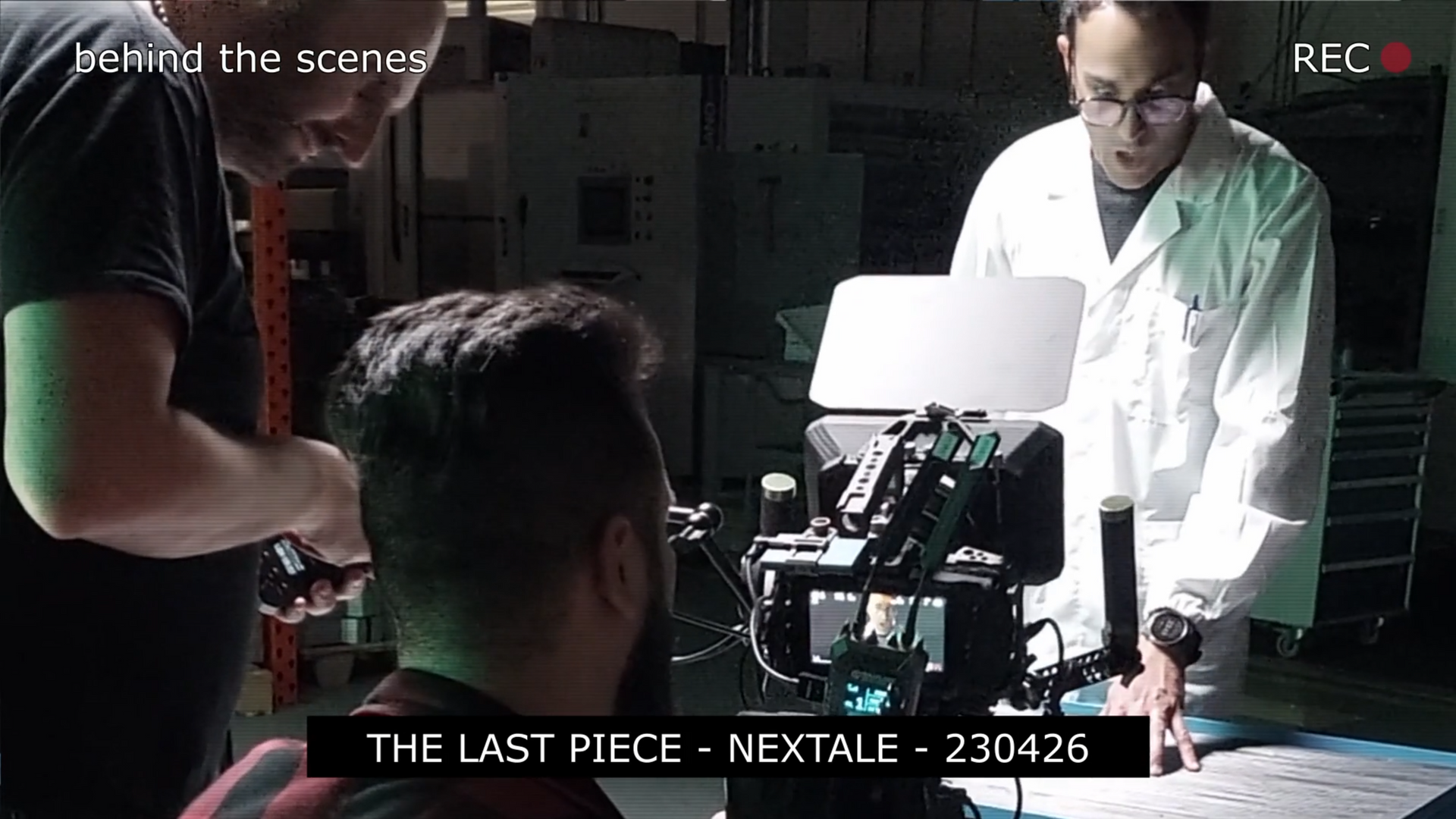 Behind-the-scenes shot. Two men setting up a camera near a person in a red shirt. White lab coat, indoor setting.