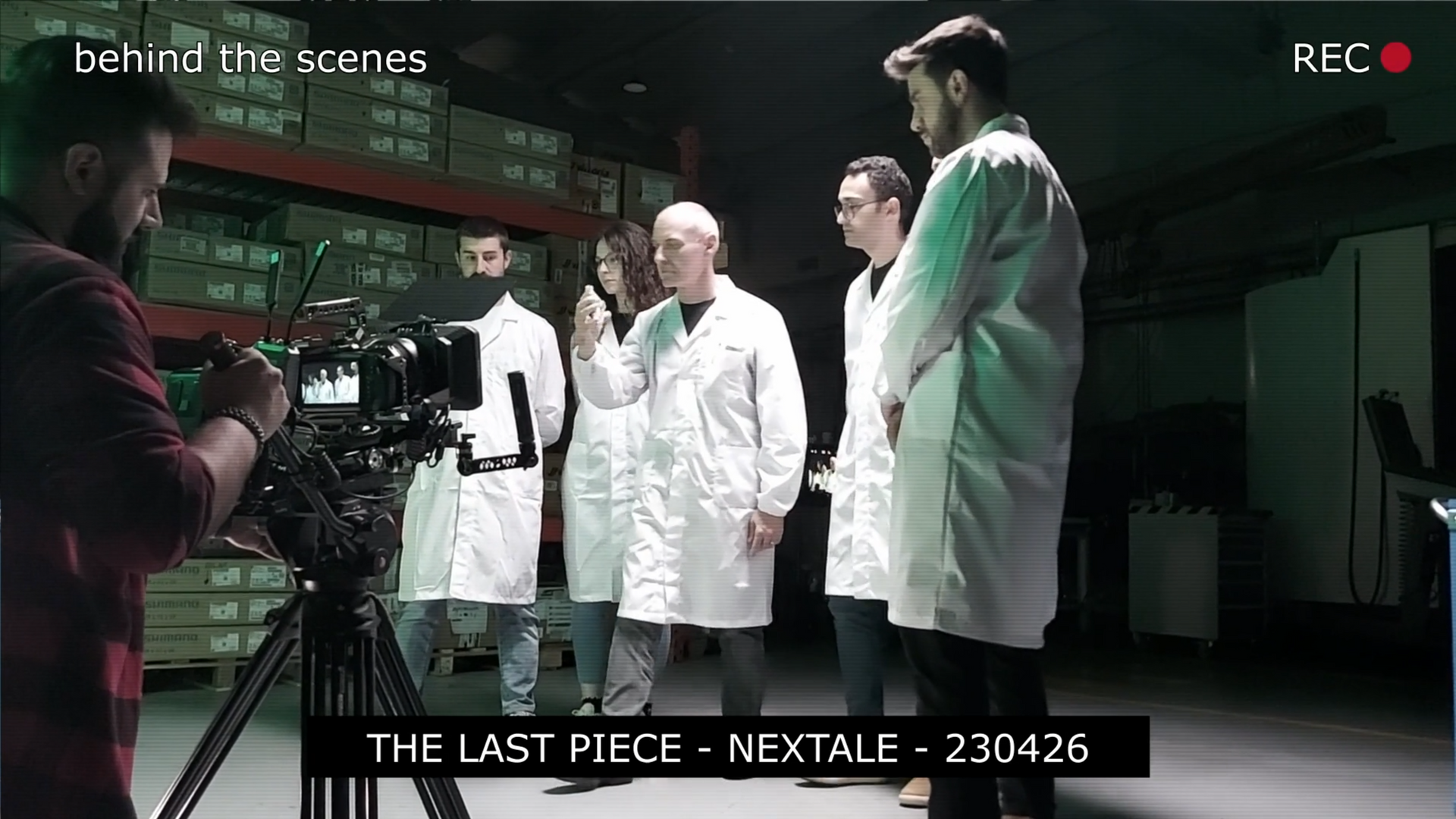 Behind-the-scenes shot of a film crew in a warehouse. Crew members wear white coats; a camera is on a tripod.