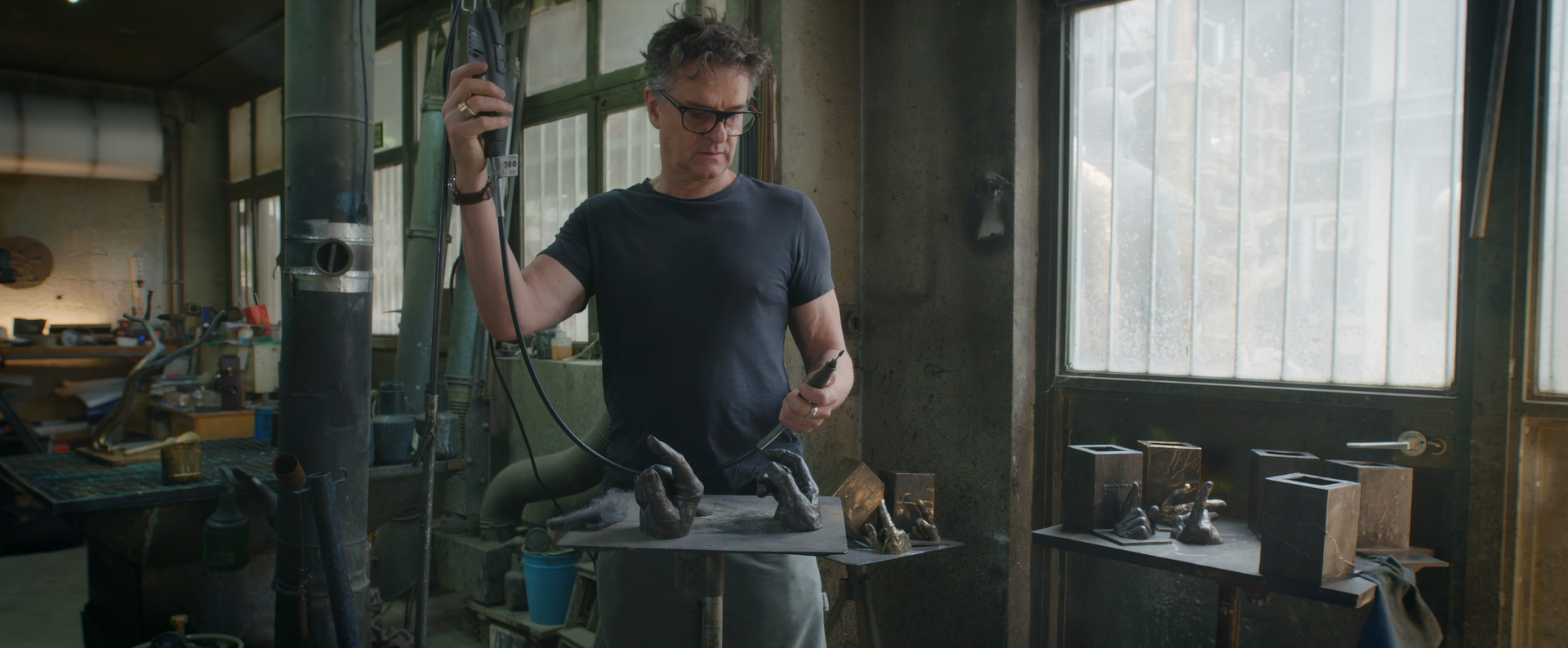 Man in workshop holds tools, examining metal pieces, near a window.