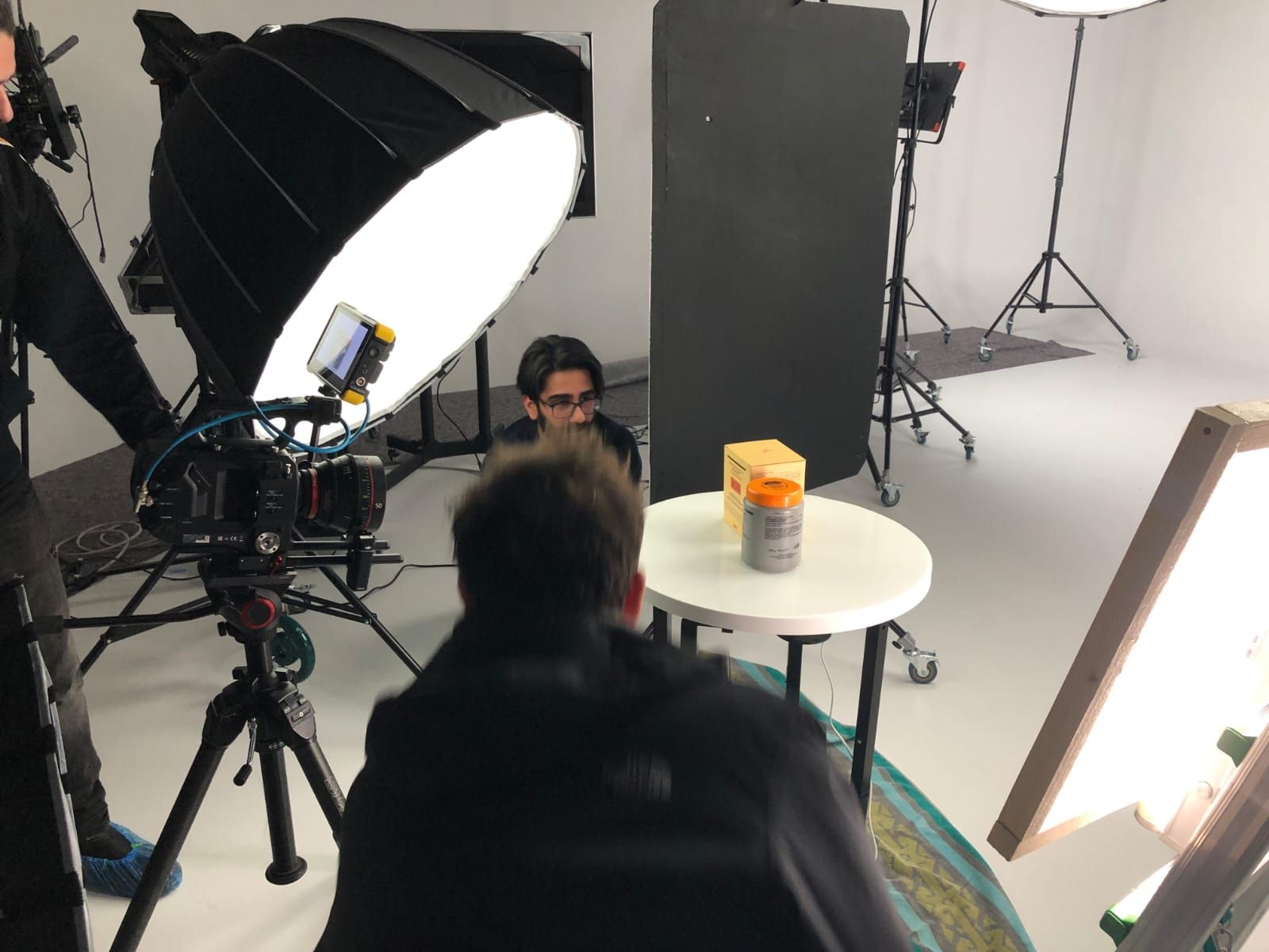 Filming a product shot: Person behind a camera, small table with a container, studio setting.