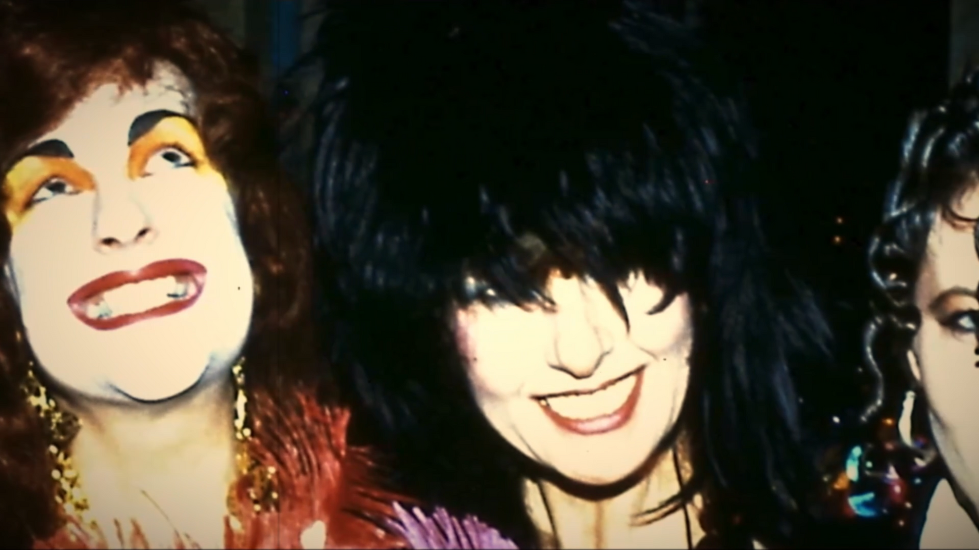 Three people with dramatic makeup and hairstyles, smiling in a dimly lit setting.