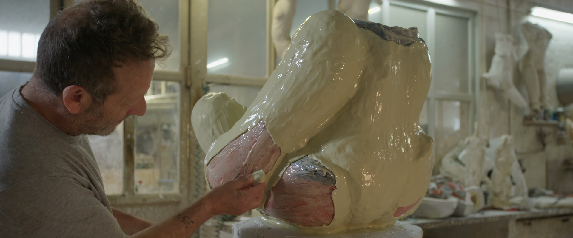 Sculptor works on a plaster torso, applying a material. Workshop setting, other sculptures visible.