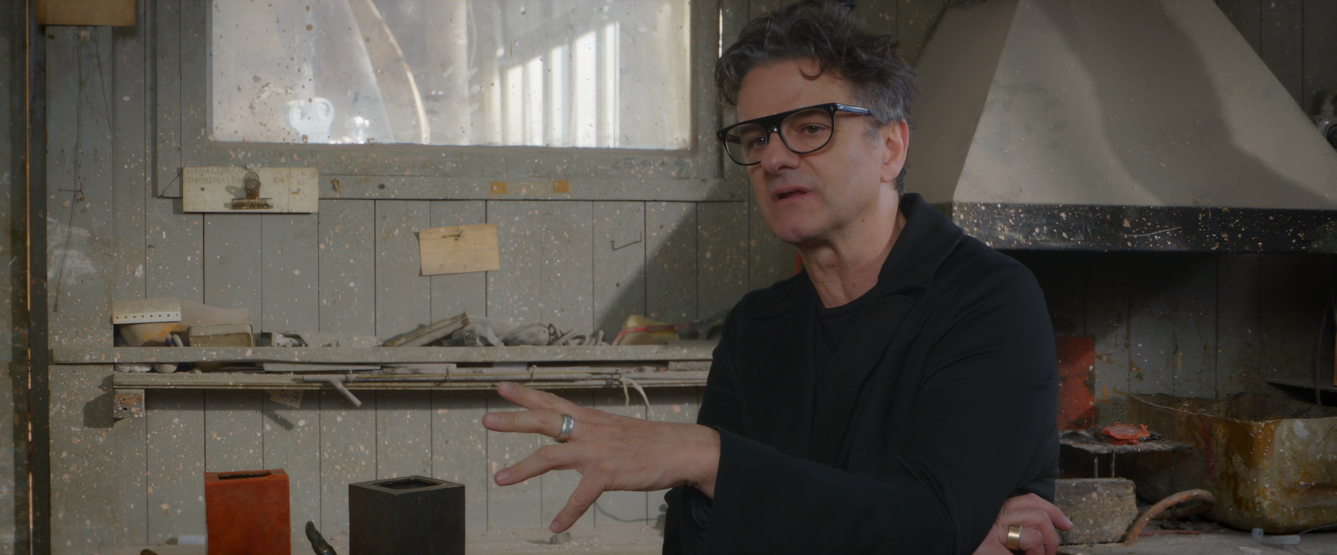 Man with glasses, gesturing with hands, in a workshop-like setting with shelves and objects.