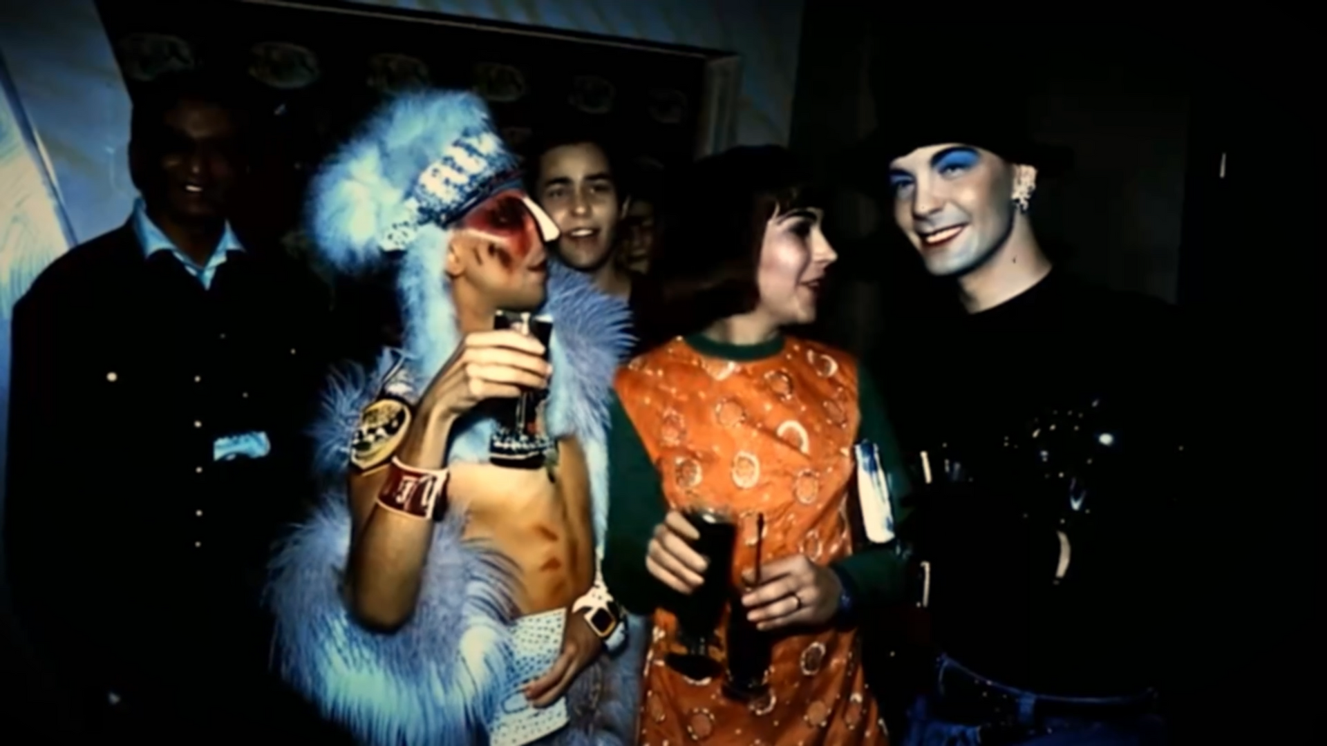 A group of people at a party: one in a headdress, others with drinks and smiling, dimly lit.