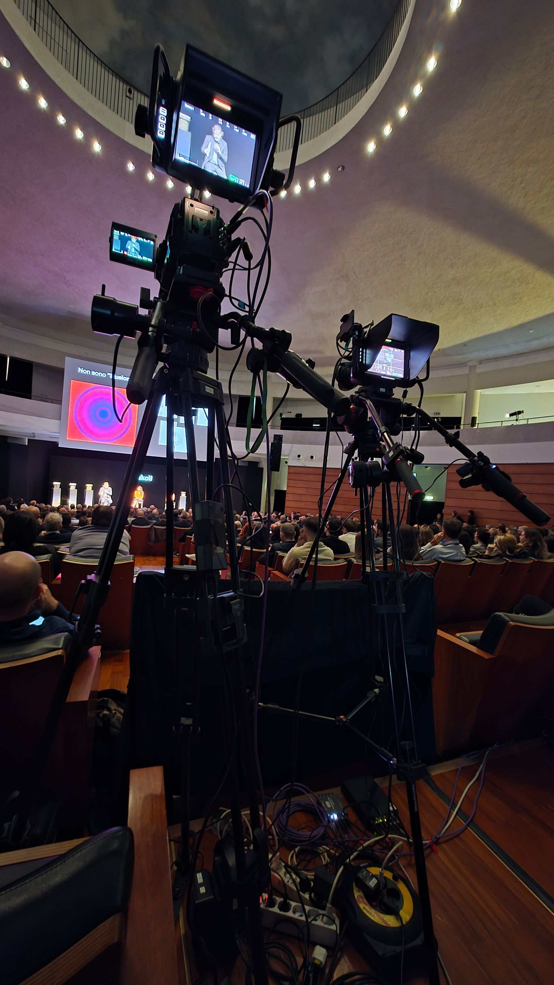 Two video cameras on tripods in an auditorium, pointed toward an event. Cables and monitors are attached.
