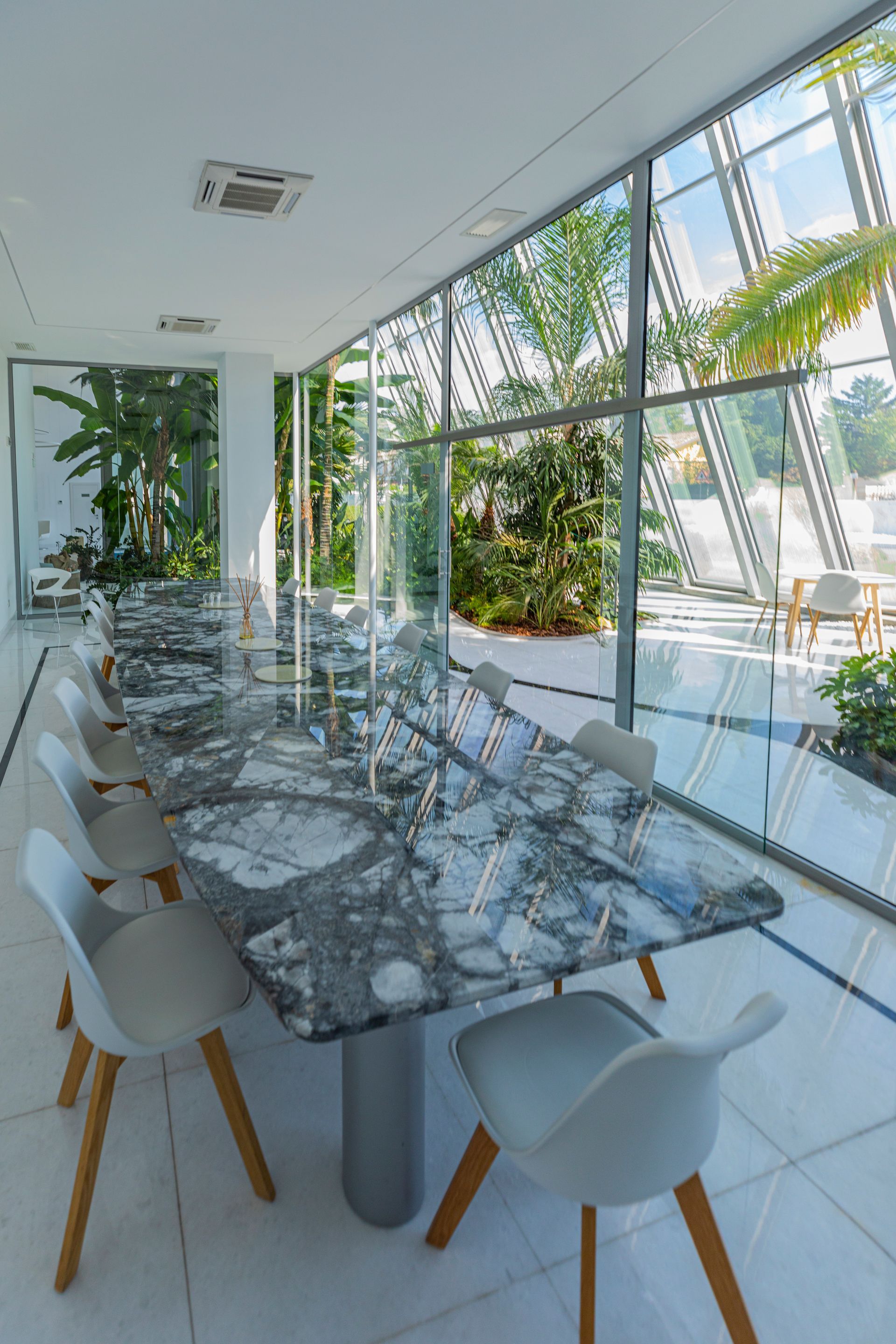Long dining table with marble top and white chairs, next to large windows overlooking greenery.