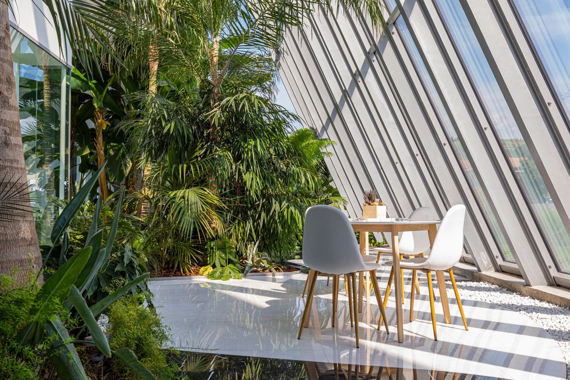 Interior with lush plants, a small table, and two white chairs, next to a large angled glass wall.
