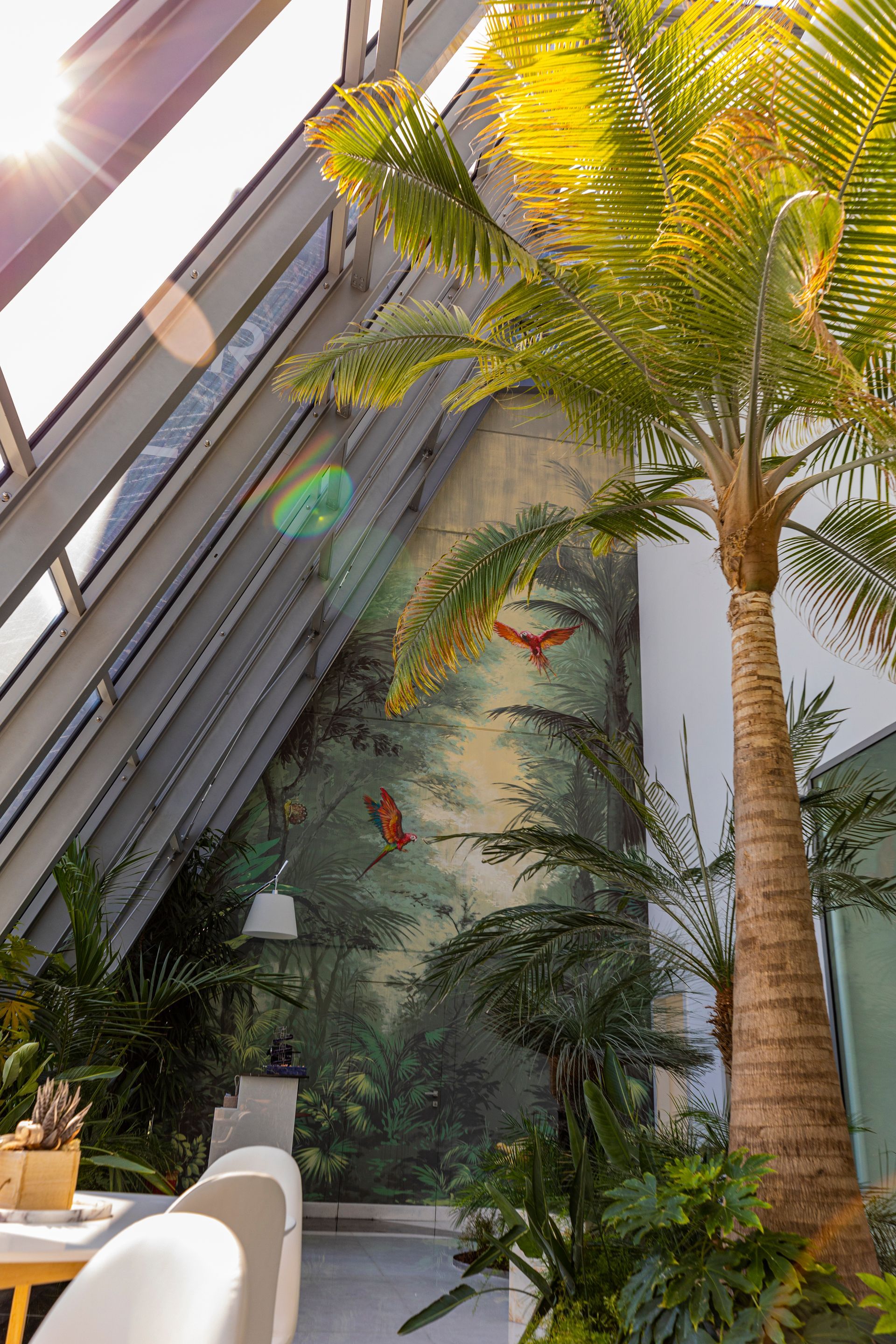 Palm tree next to a mural of a jungle scene with two red birds. Sunlight shines through the glass ceiling.