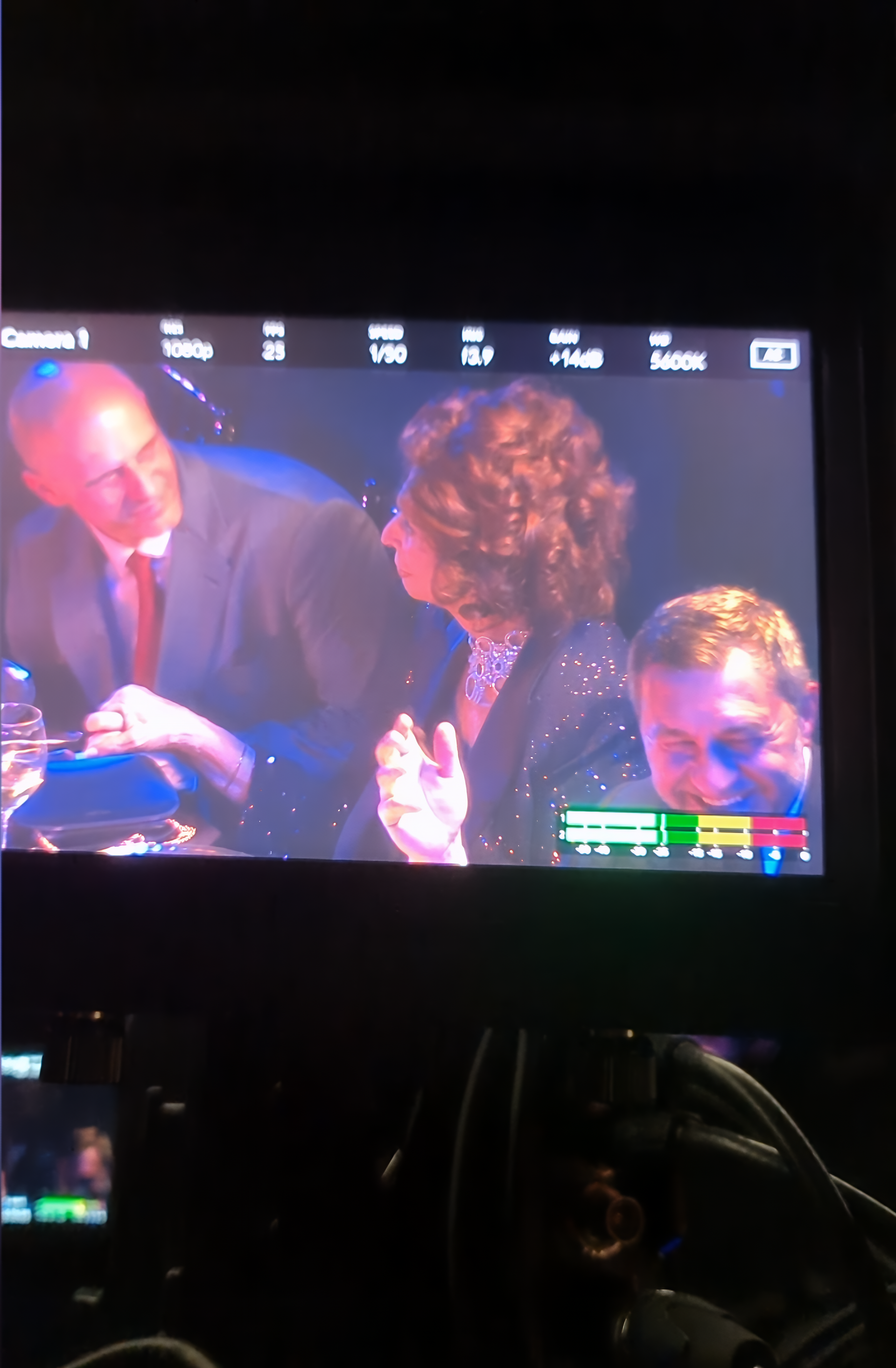Screen displaying people at a formal event; woman applauds, man gestures, another man smiles.