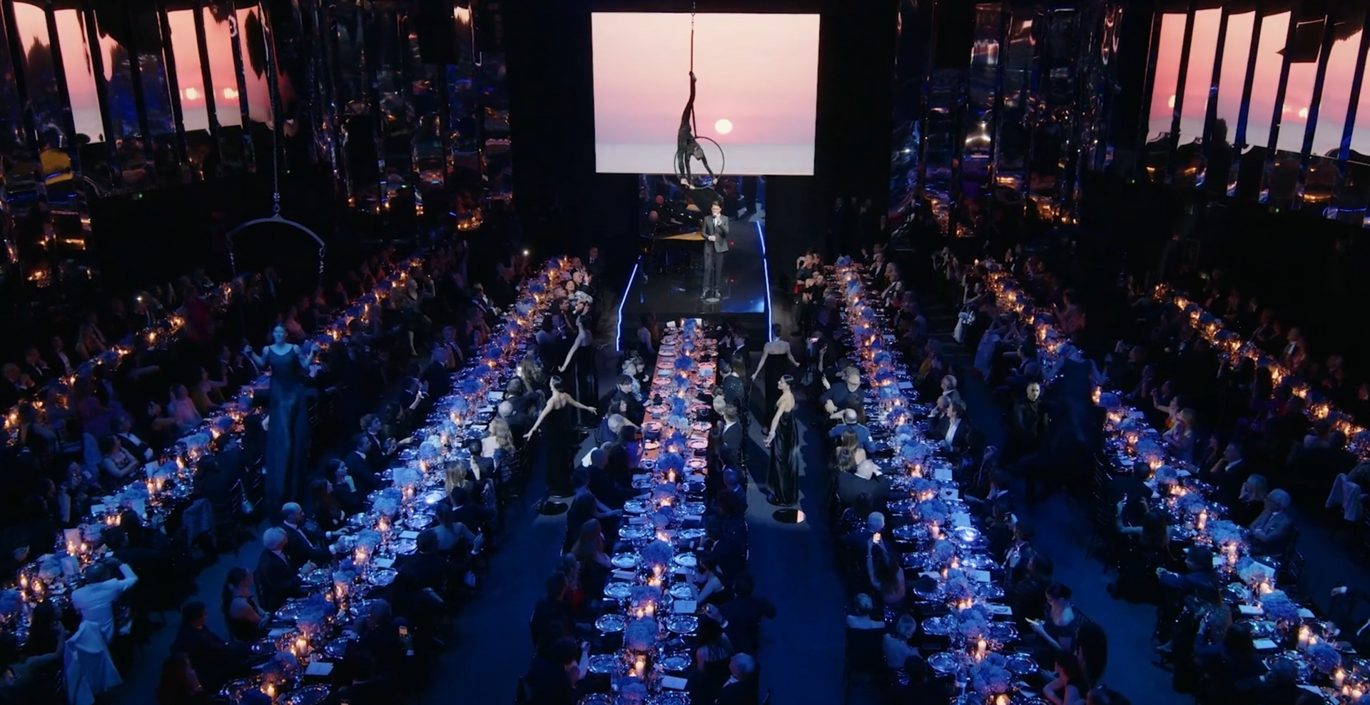 Aerialist performs above a seated gala. Long tables lit by candles, projected sunset backdrop.