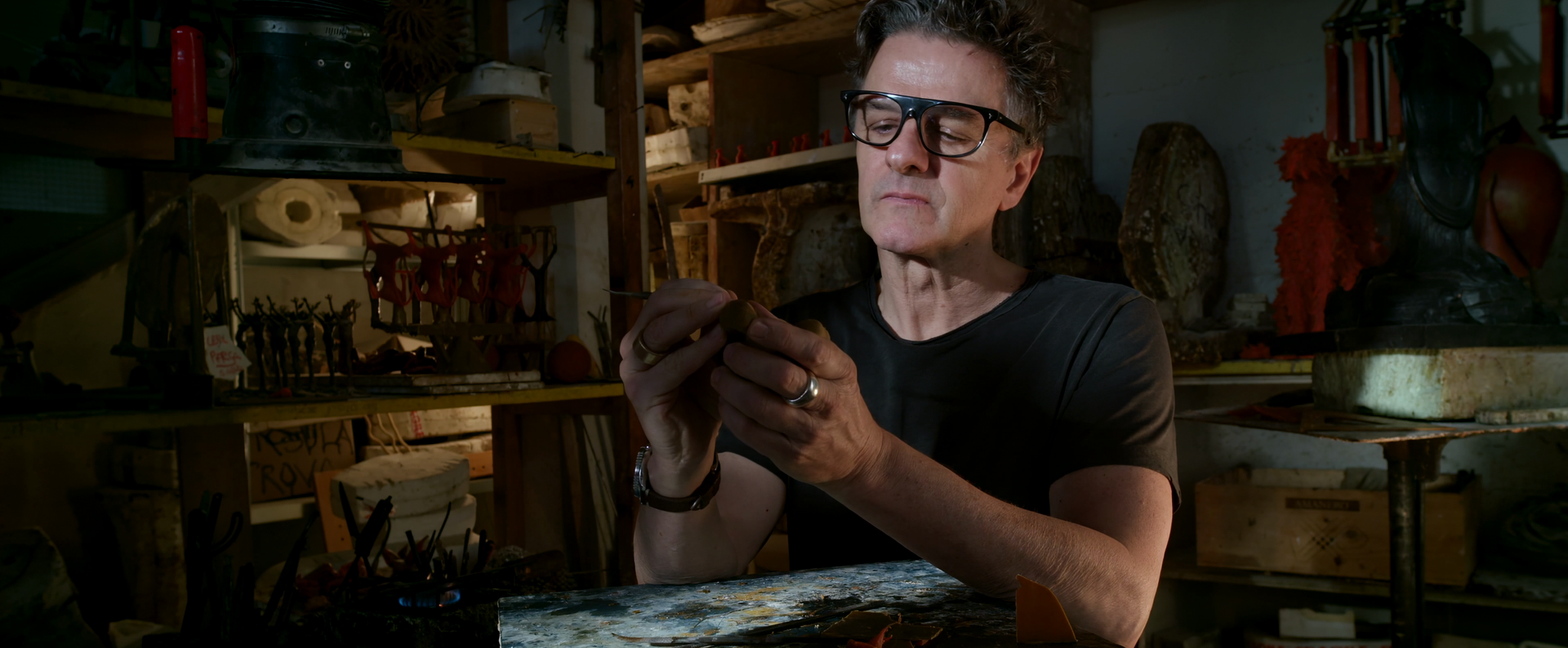Man with glasses in a workshop, looking at object in his hands. Shelves and tools in the background.