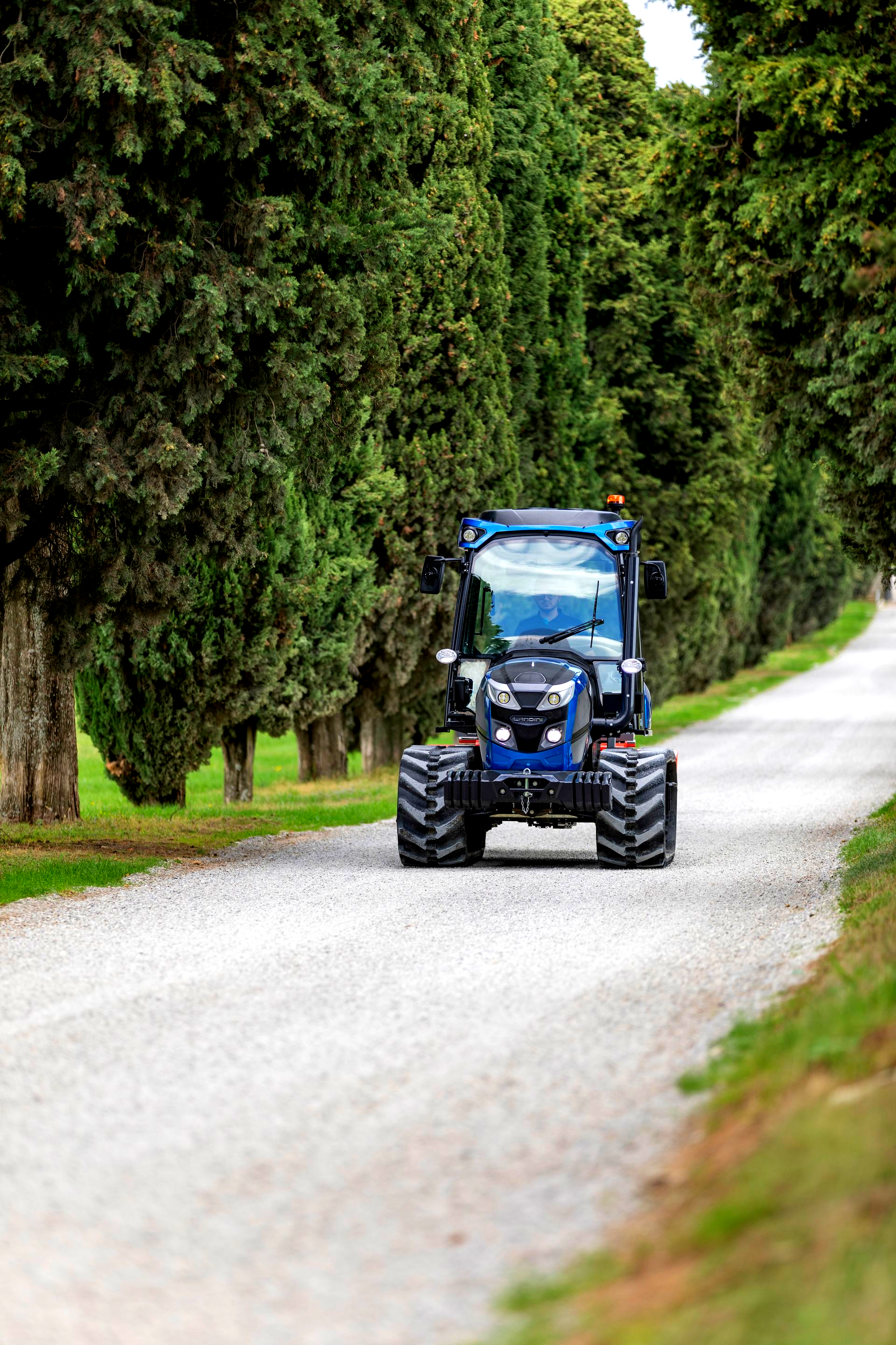 Blue tractor driving on a gravel road lined by tall green trees.