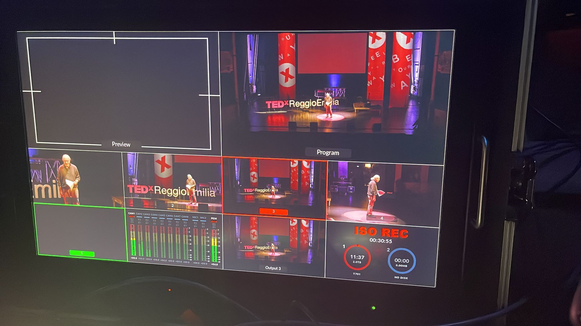 Live event multi-camera view on a monitor, displaying speakers on stage with red and white decor.