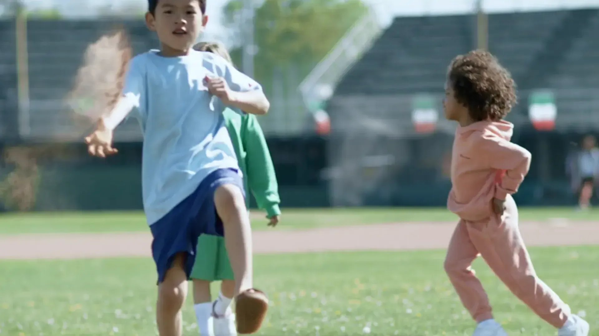 Children playing on grass field; boy kicking up dirt, two others running nearby.