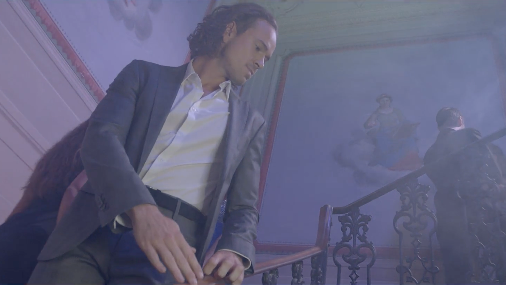 Un uomo in giacca e cravatta guarda in basso verso le scale, illuminate da una luce blu e soffusa.