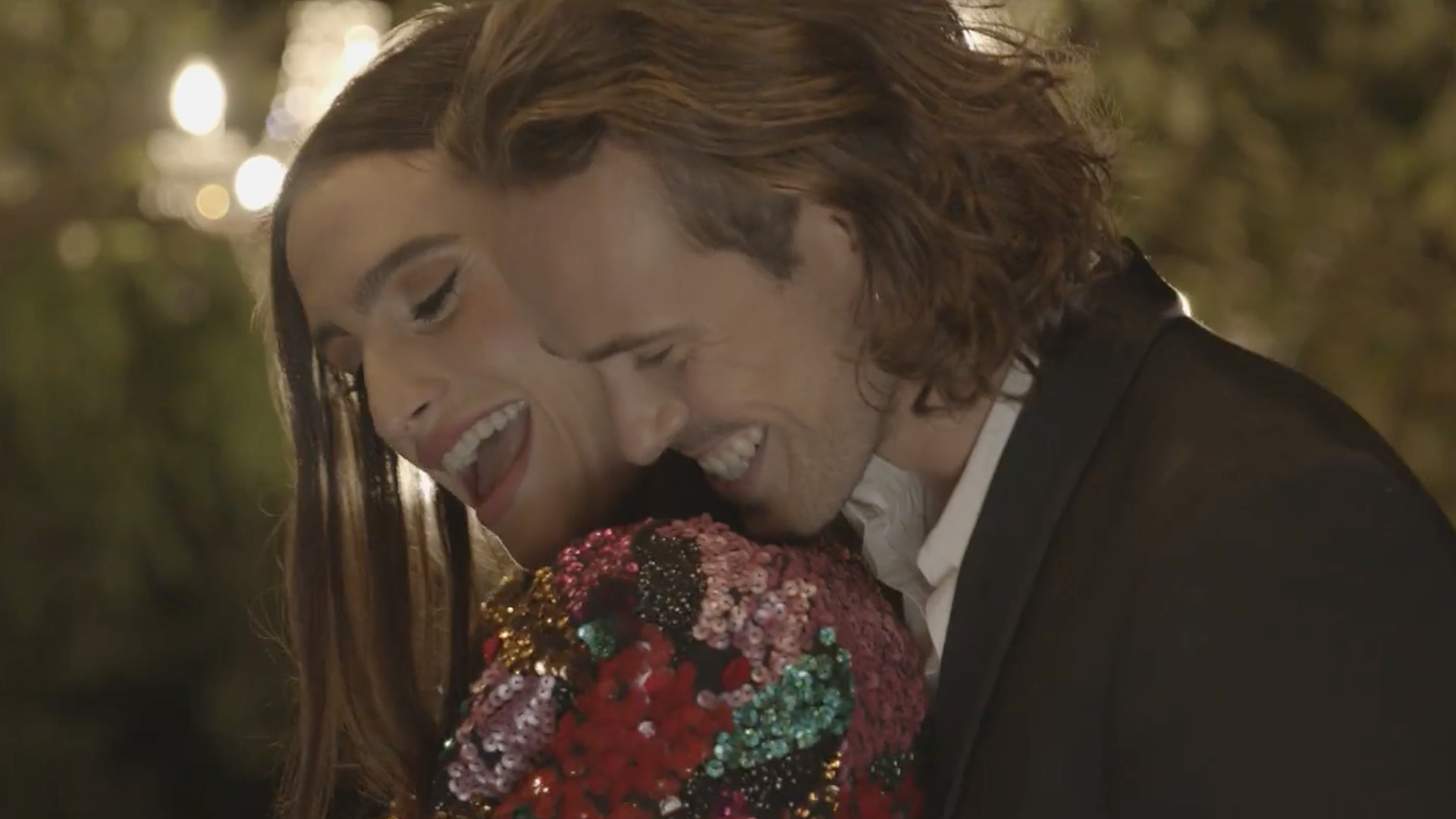 Una coppia si abbraccia e ride, la donna indossa un top con paillettes, durante un evento serale all'aperto.