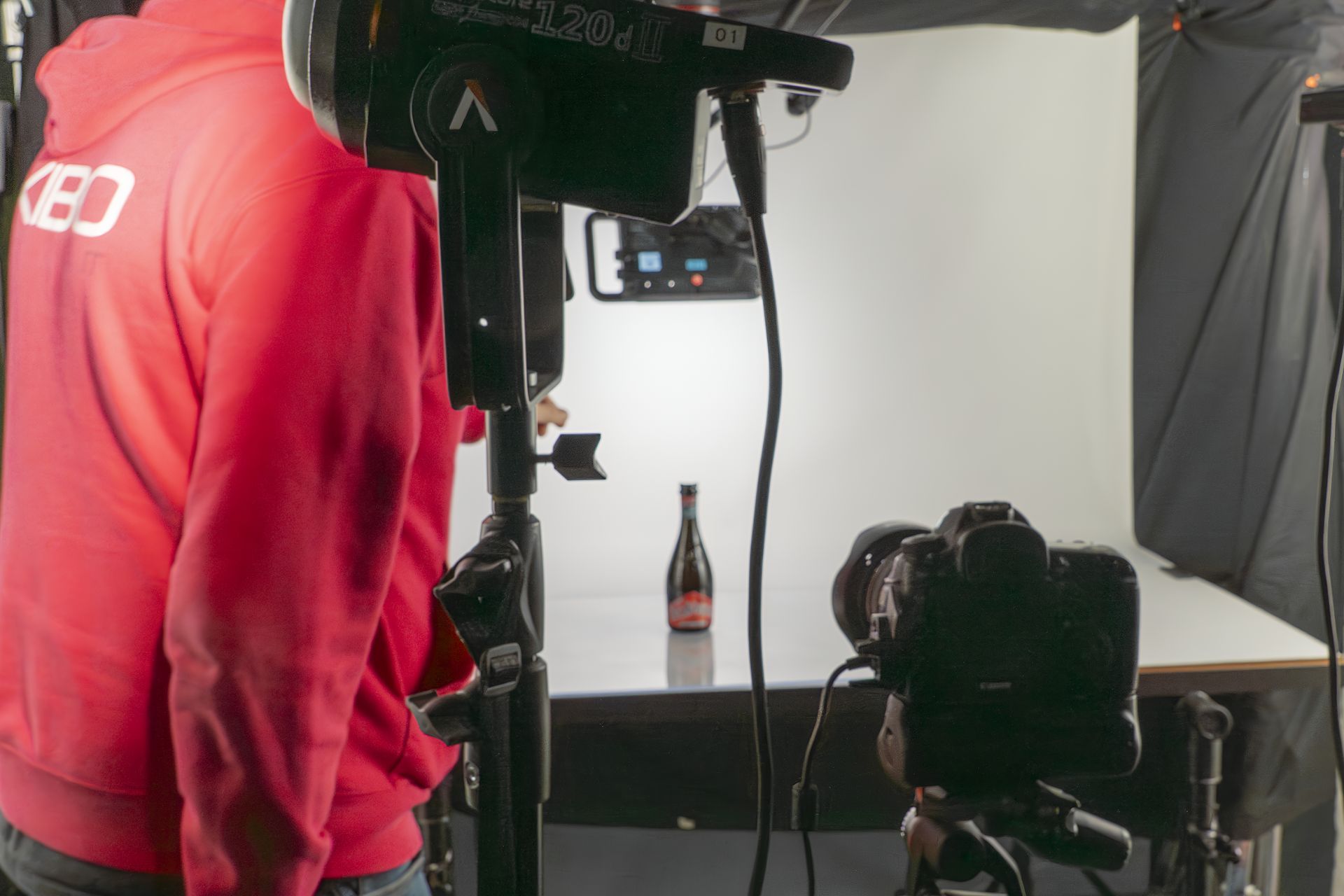 Photographer in red hoodie sets up studio lights, camera, and bottle on table against white backdrop.