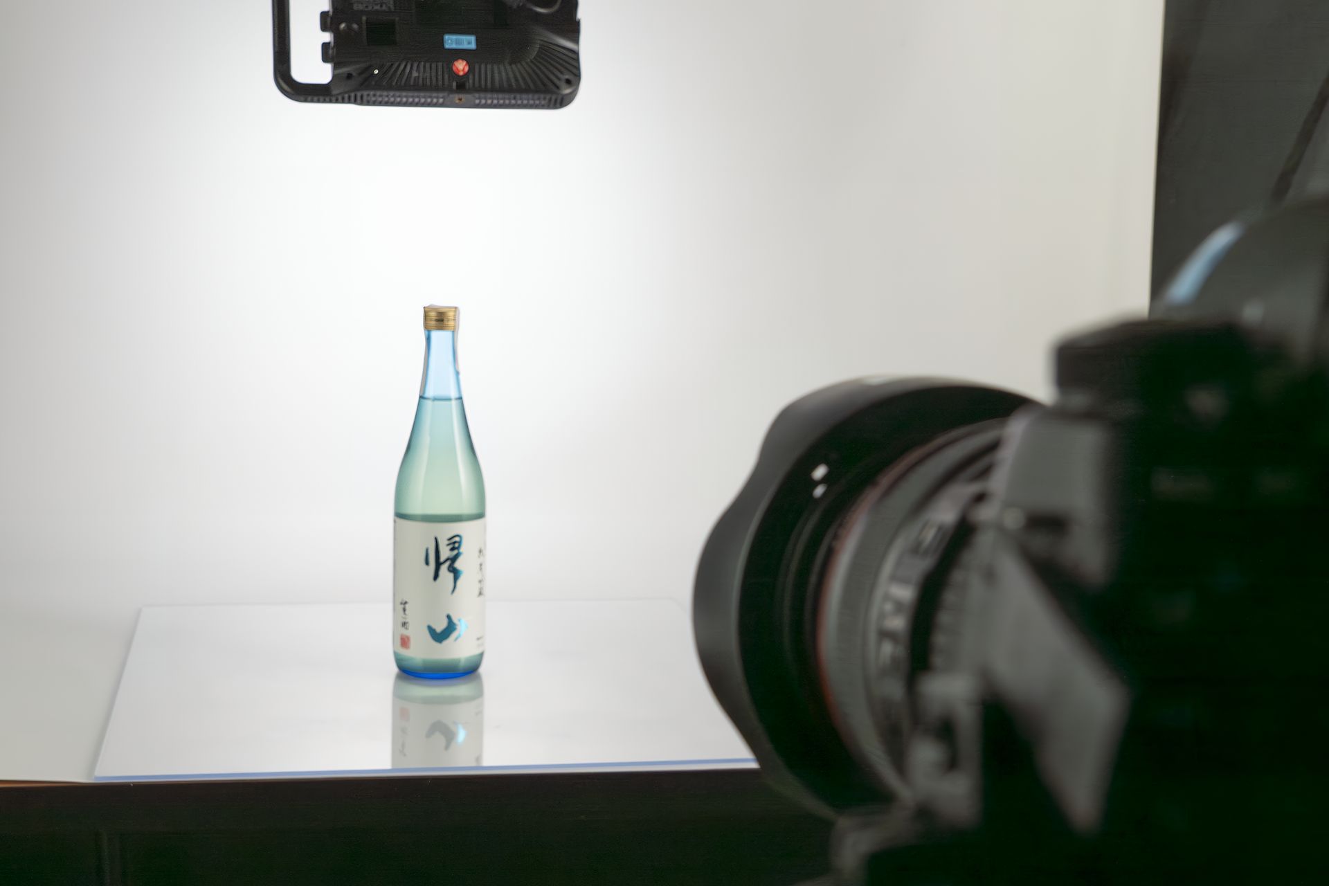 Sake bottle on reflective surface, photographed with camera and overhead light against a white backdrop.
