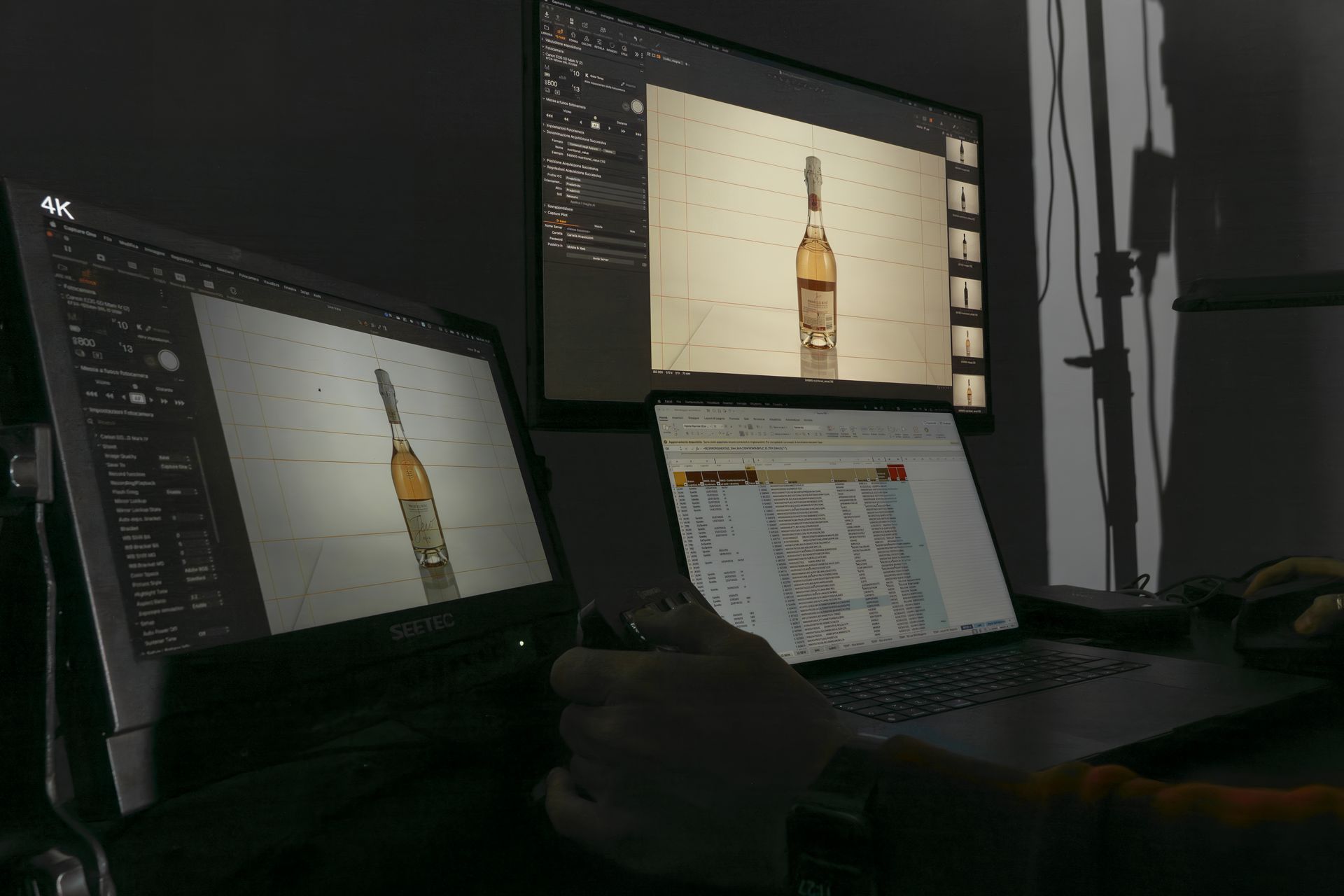 Person working on a laptop and two monitors displaying product photography; a bottle with a tan background.