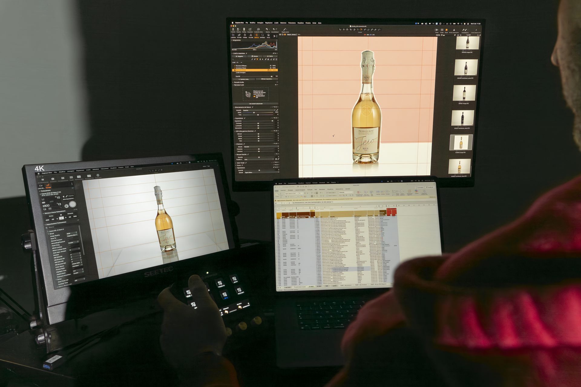 Person edits a photo of a bottle on multiple screens using a tablet and laptop in a dimly lit setting.
