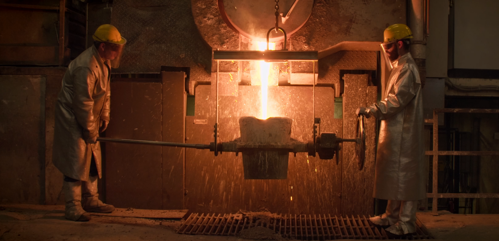 Two workers in protective gear pour molten metal in a factory. Sparks fly in the orange glow.