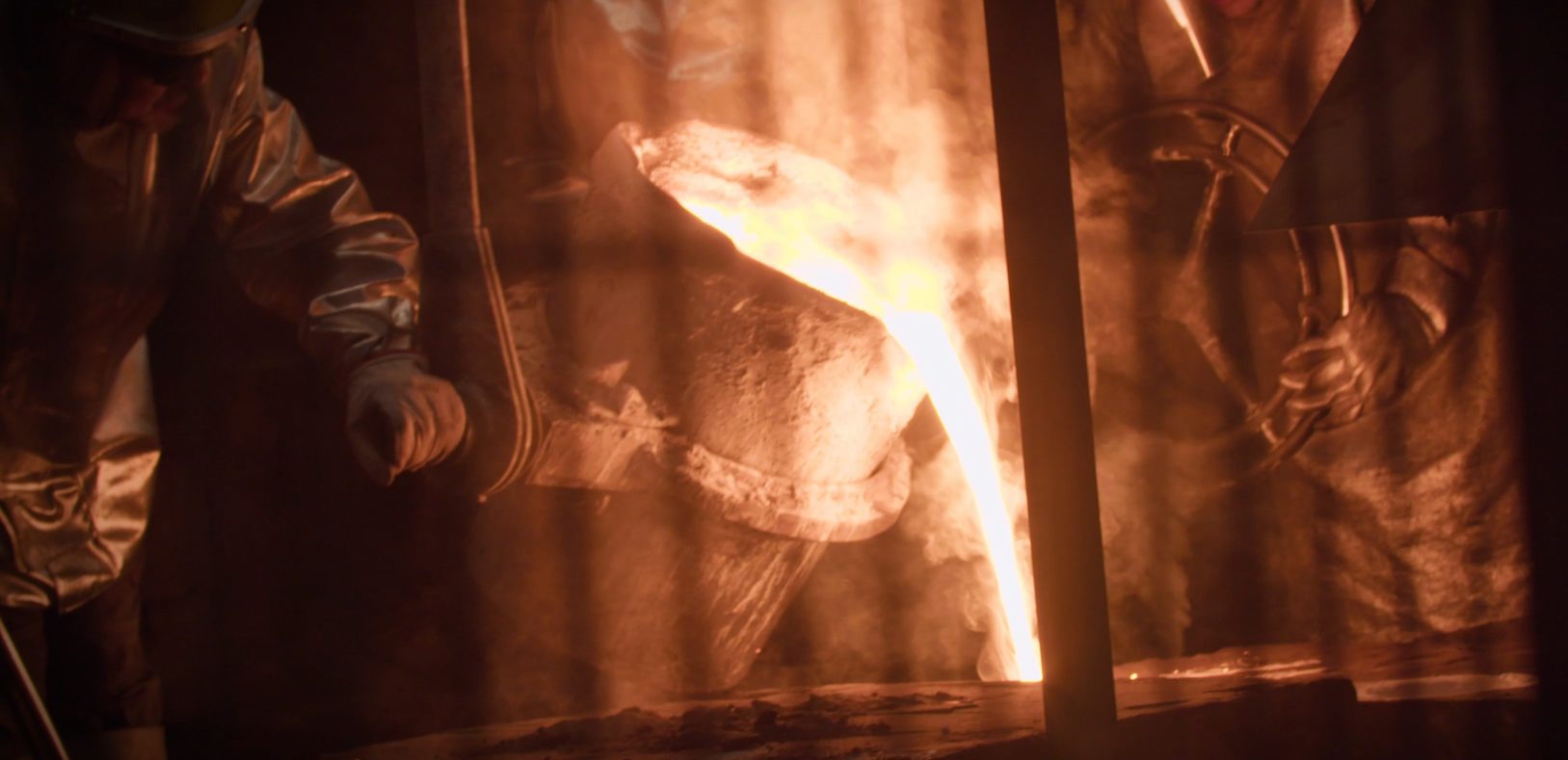 Molten metal pouring from a container in a factory. Workers in protective gear handle the hot liquid.