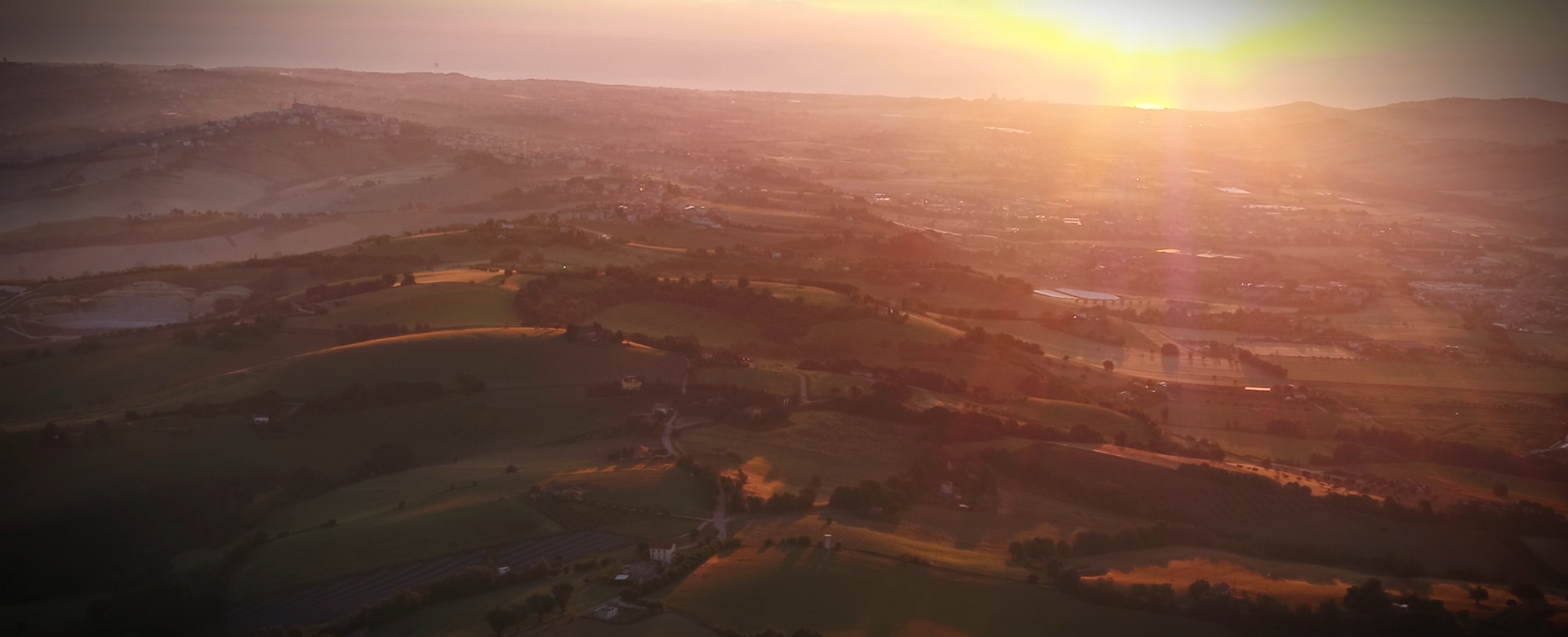 Aerial view of a landscape at sunset. Orange sunlight illuminates hills and valleys with a hazy atmosphere.