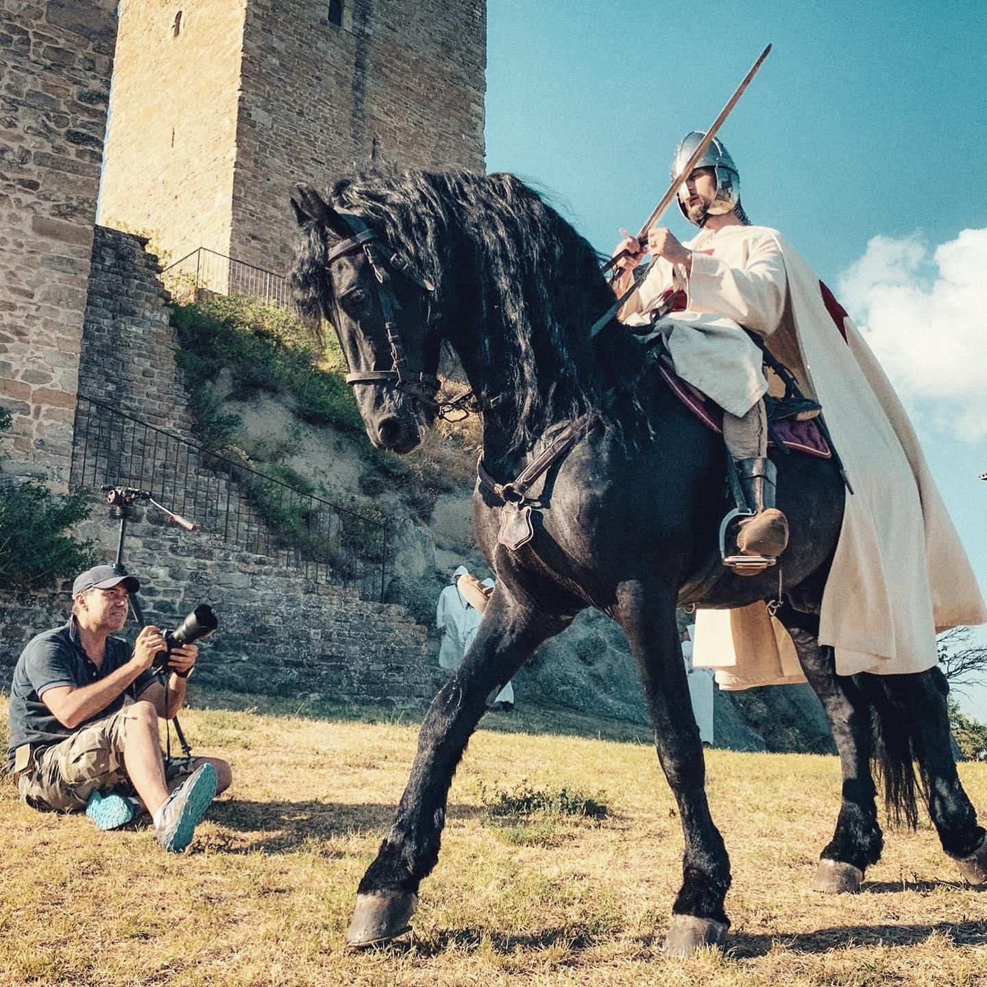 Knight on black horse, holding spear, near stone building, photographer taking photos.