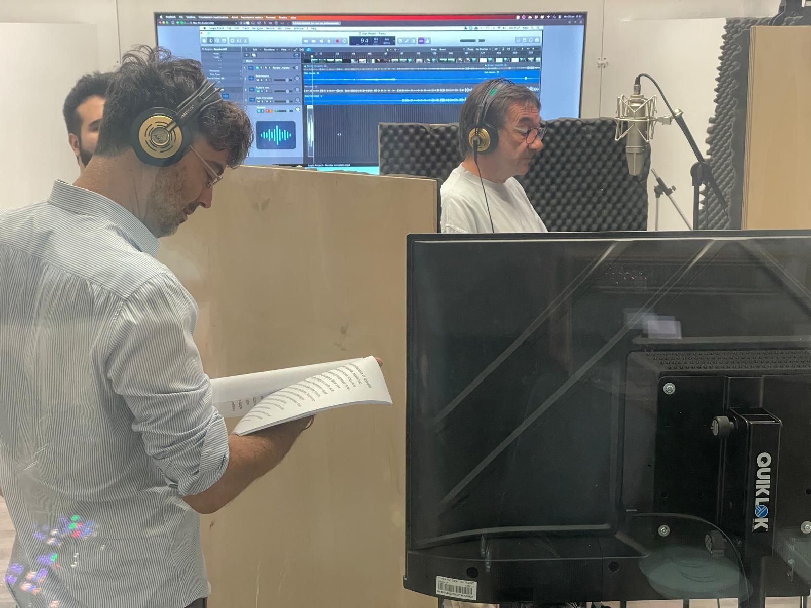 Three men in a recording studio; one reading script, two wearing headphones near a microphone.