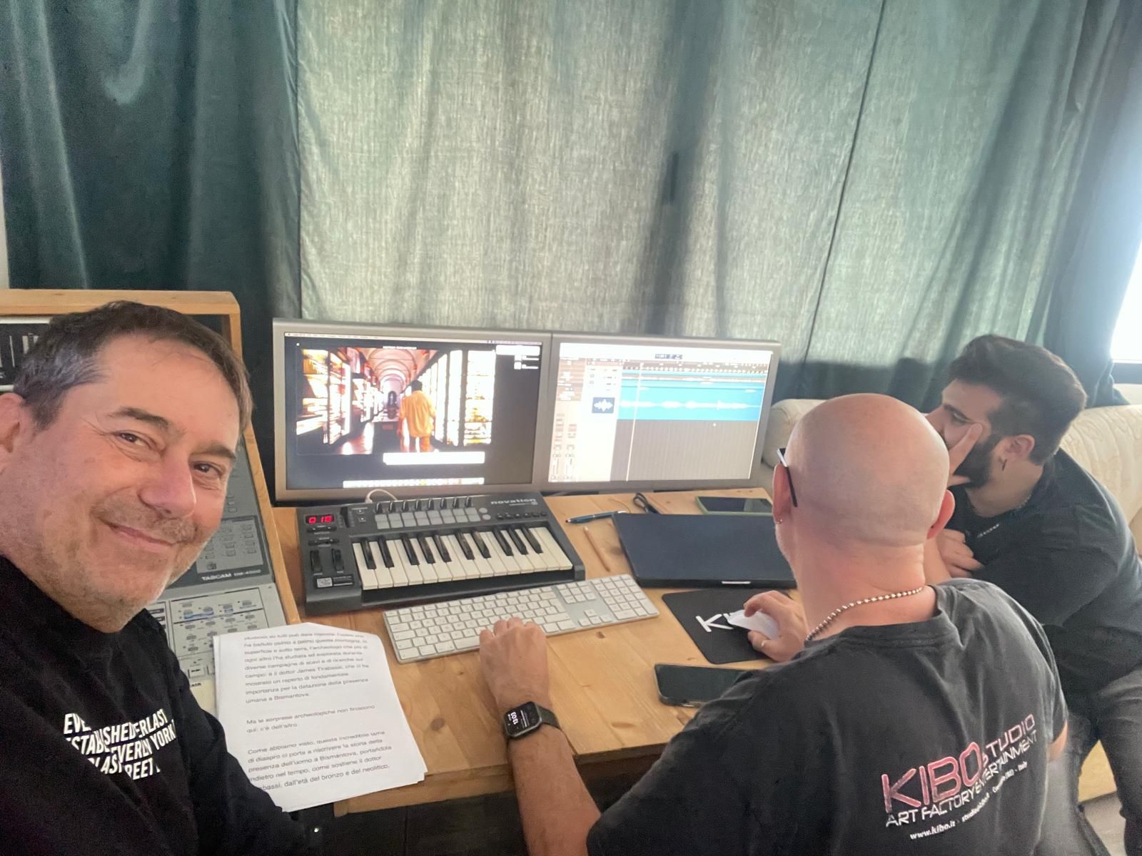 Three people at a desk in a studio. One smiles, two are at computers, editing. Sunlight enters room.