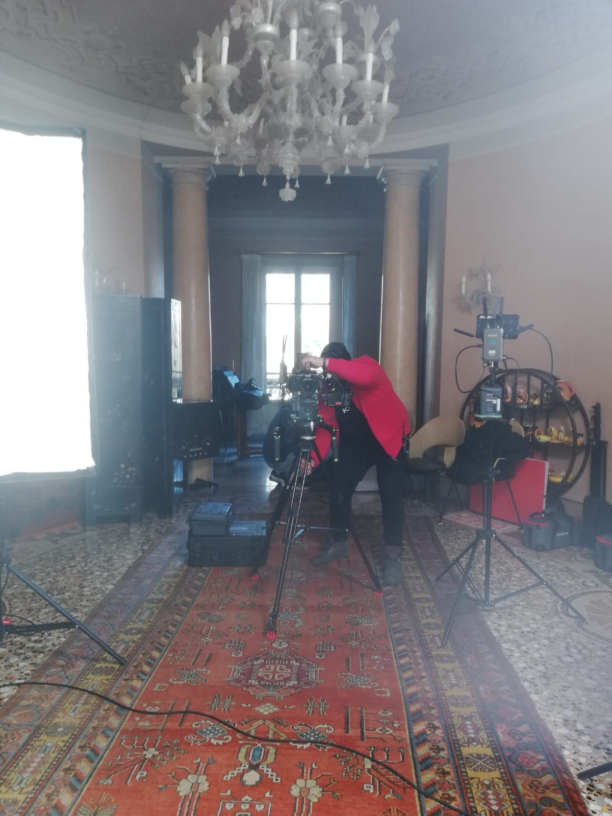 A person in red jacket filming with a tripod-mounted camera in an ornate room with a chandelier.