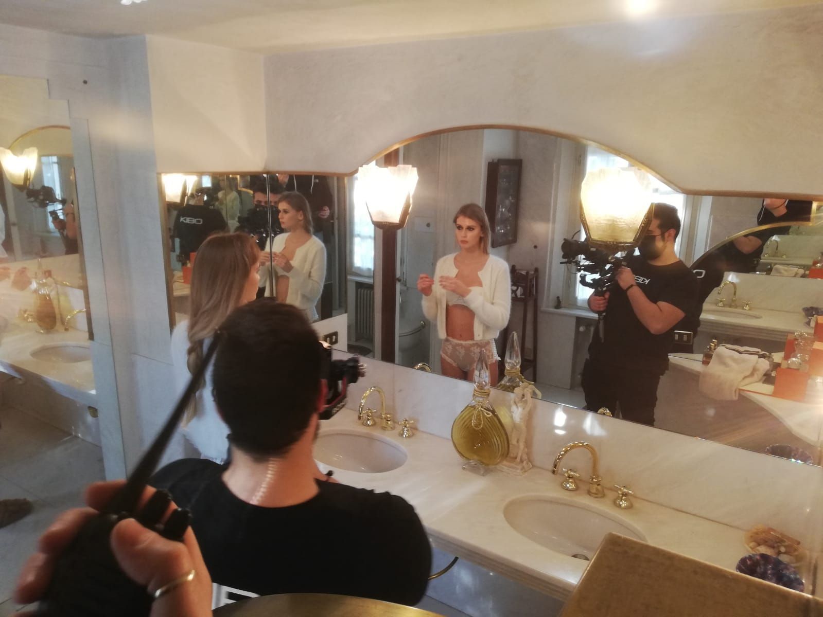 A model being filmed in a bathroom with a camera crew. She wears lingerie, mirror reflects the scene.