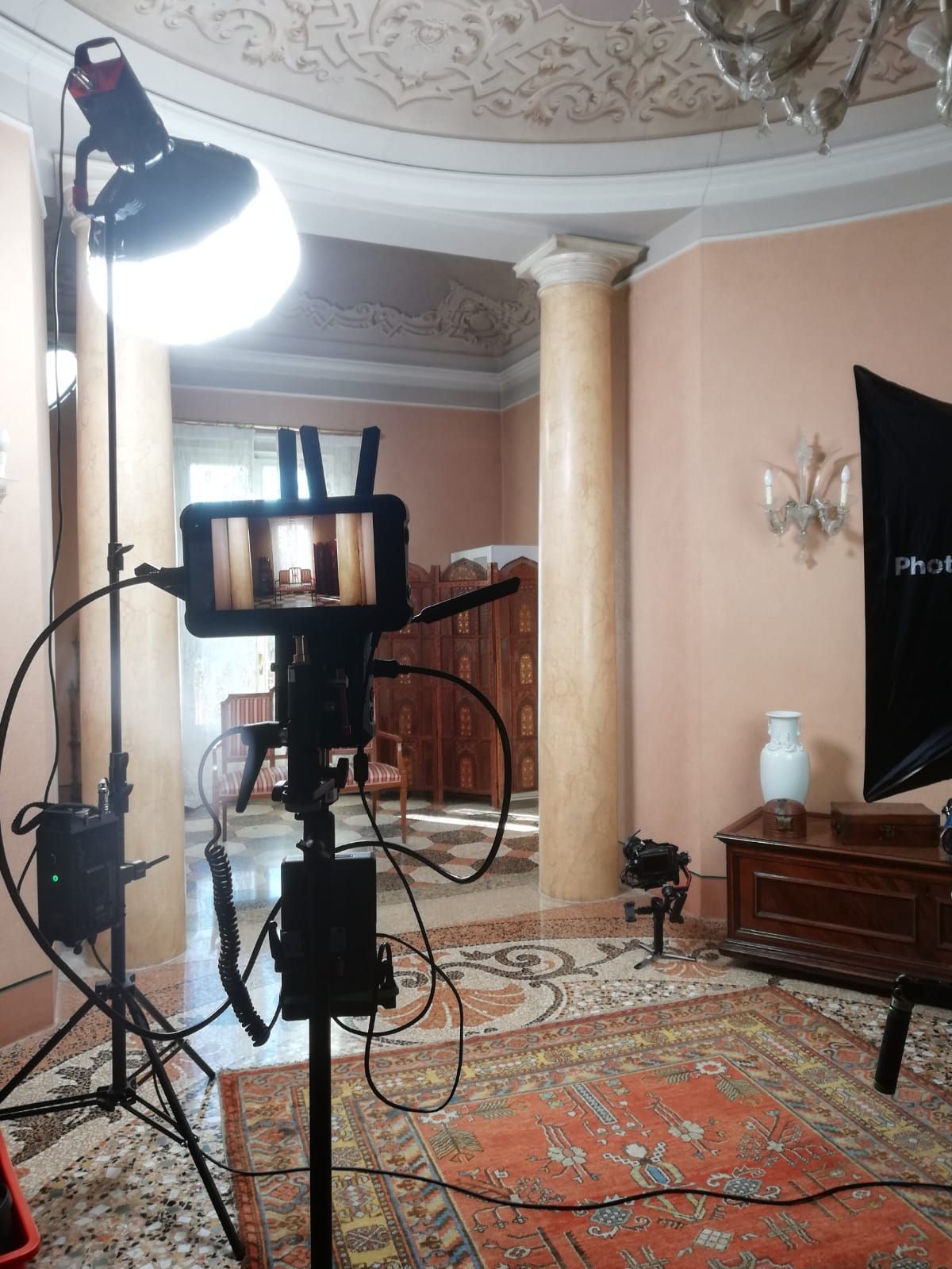 Film set: a phone camera on a tripod, lights, and equipment in an ornate room with a rug and columns.