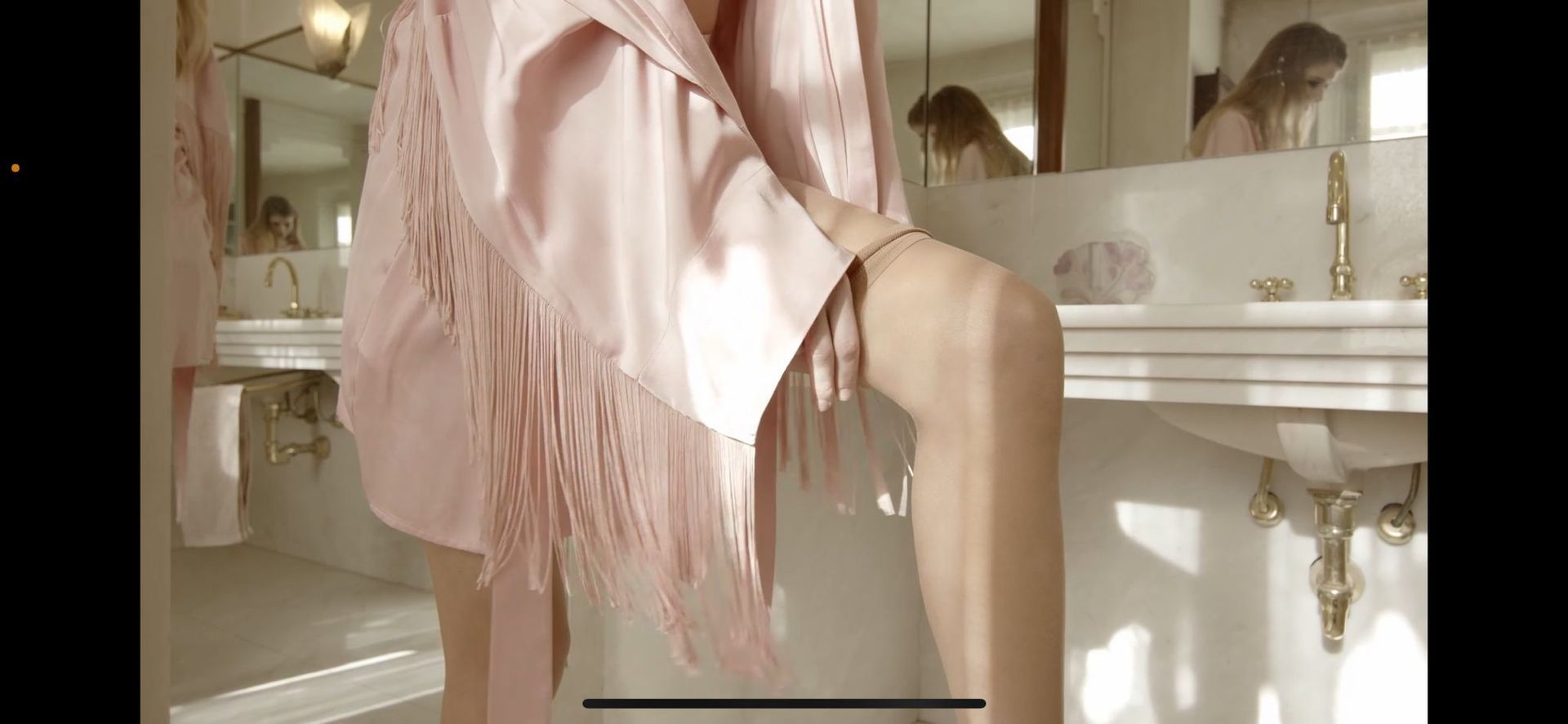 Person in a pink robe touching their leg in a bathroom. There are reflections of a person in the mirror.