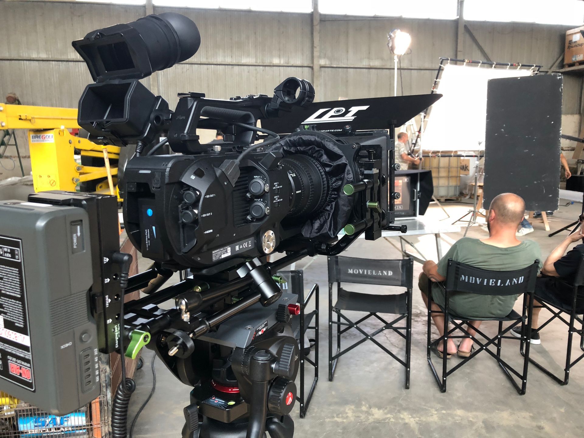 Large film camera on tripod, pointed towards a person seated in a director's chair in a studio.